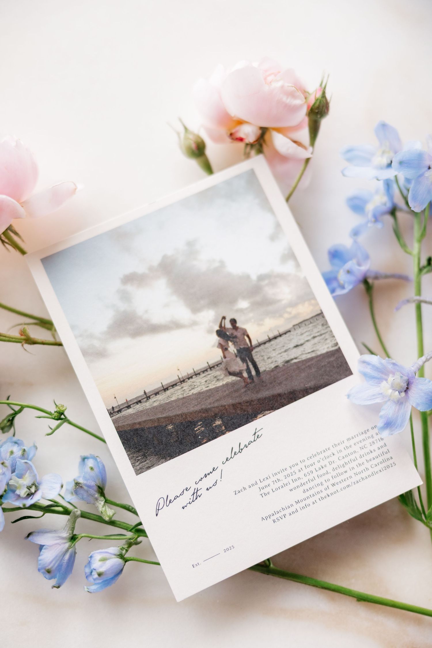 A romantic photograph surrounded by pink roses and blue flowers showing a couple silhouetted against a beach sunset.
