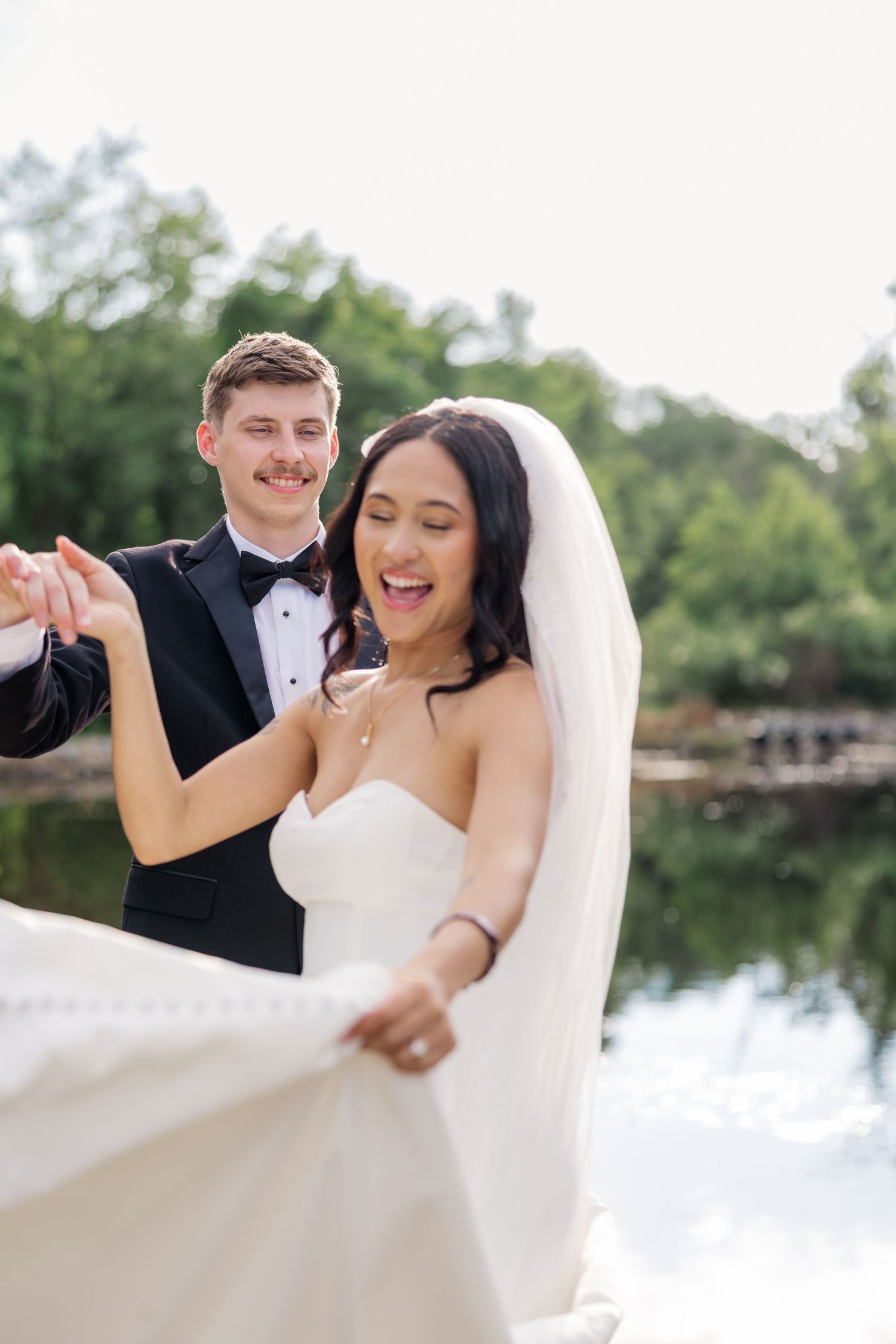 Joyful couple in wedding attire dancing and twirling by a reflective lake.