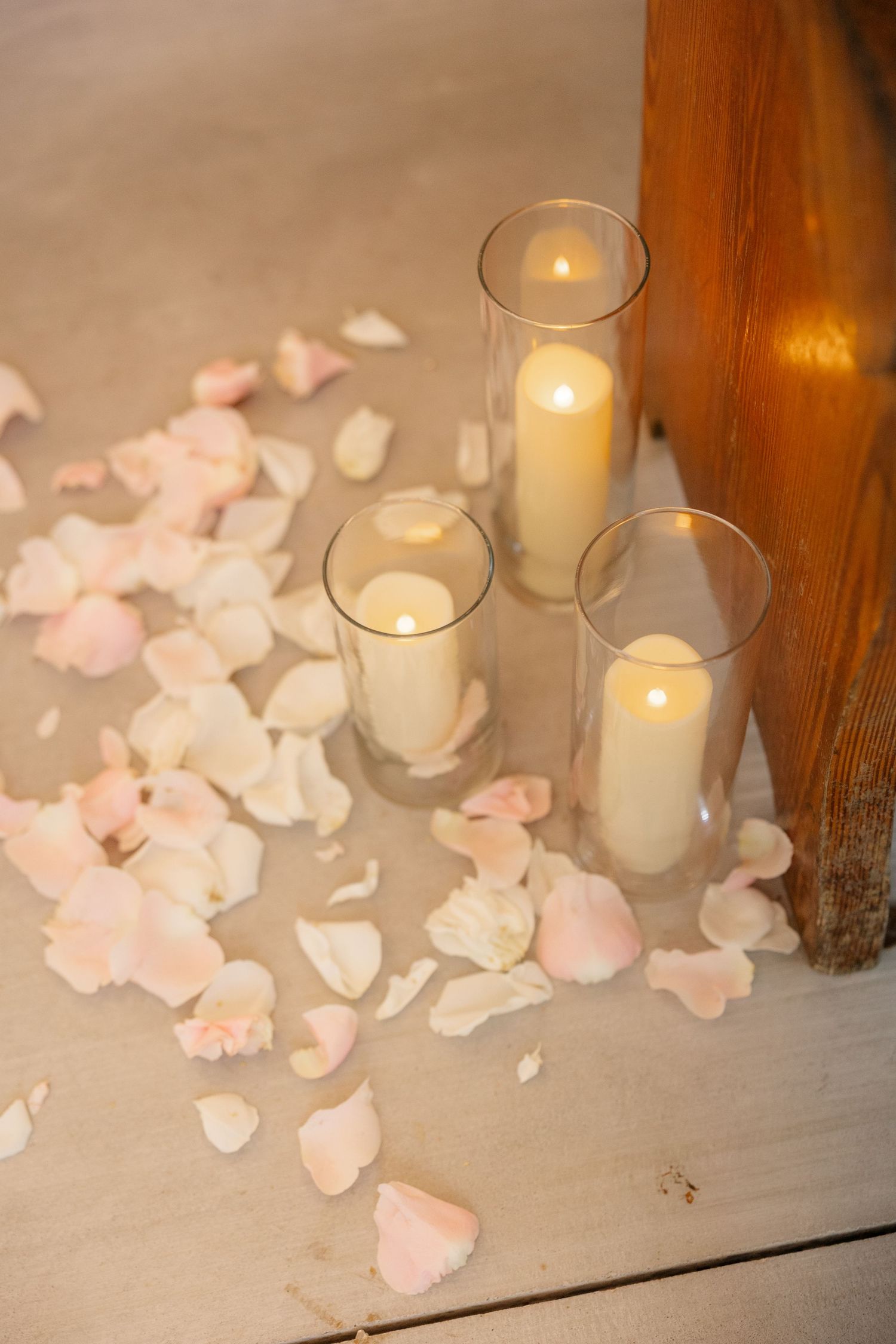 White candles in glass holders with scattered pink rose petals creating romantic wedding decor.