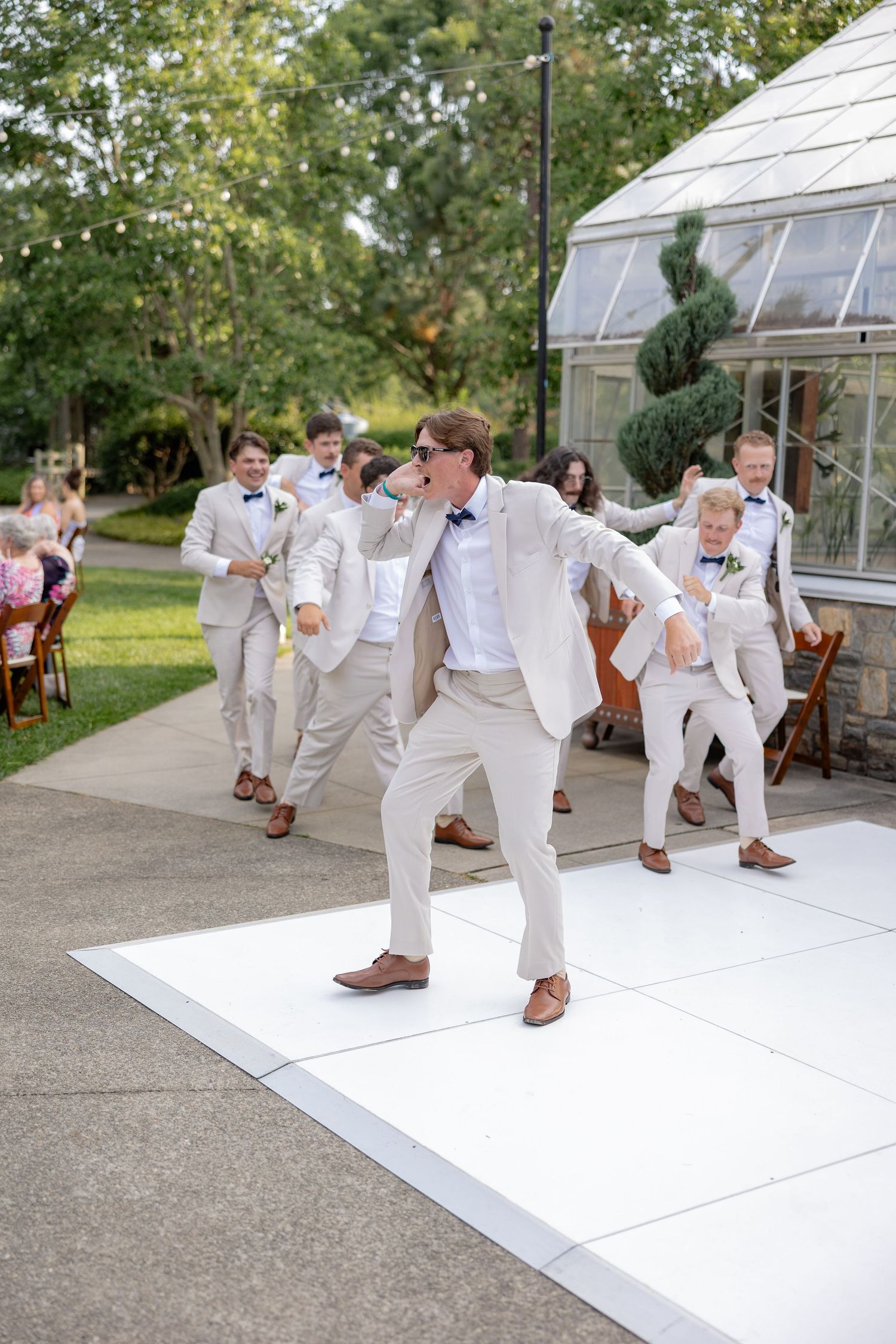 Groomsmen in light colored suits dance and celebrate on a white dance floor outside a greenhouse venue.