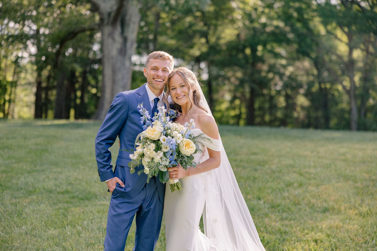 Bride and groom posing together in lush green garden with yellow and blue bridal bouquet.