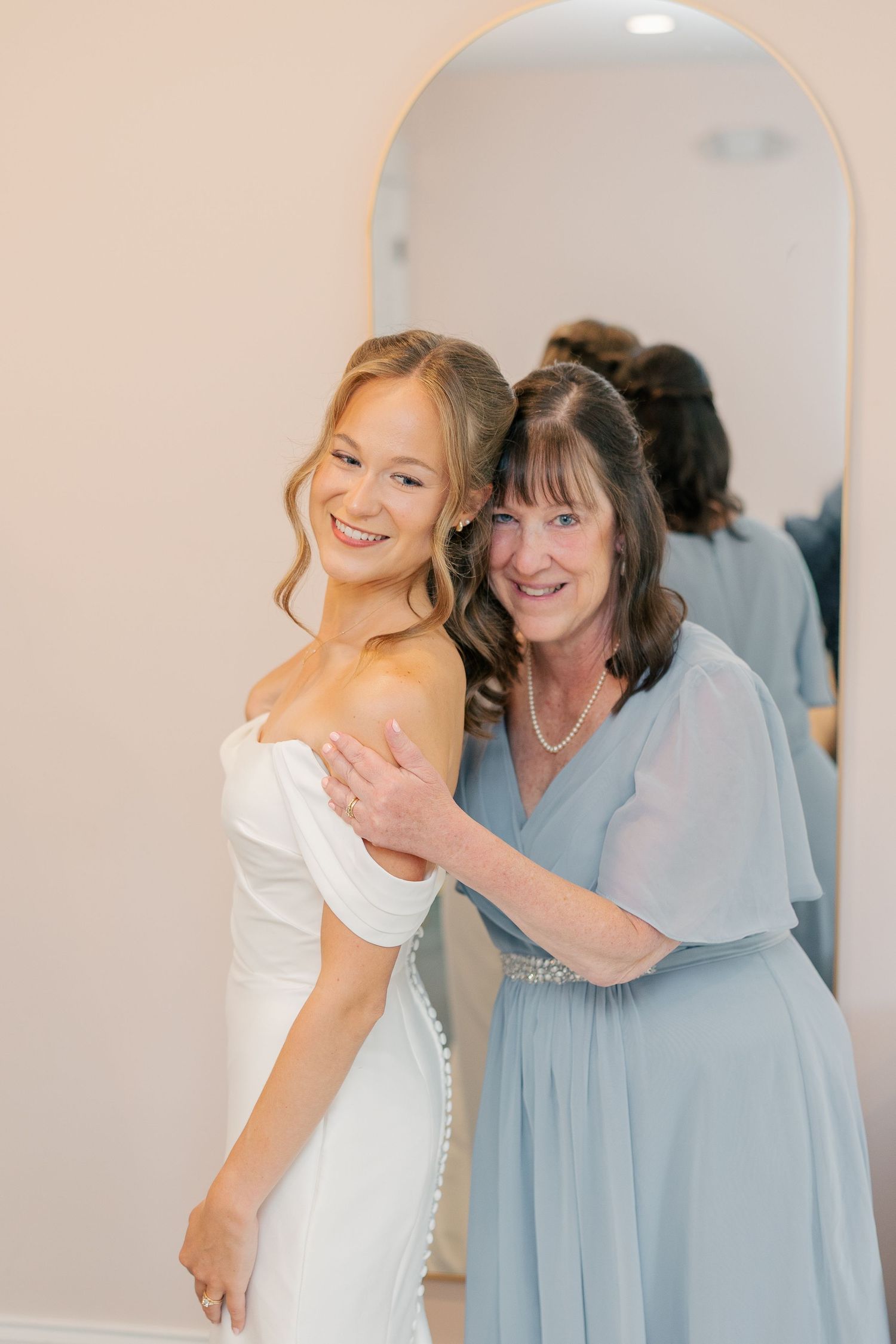 Smiling bride in off-shoulder white gown poses with older woman in blue dress in front of arched mirror.