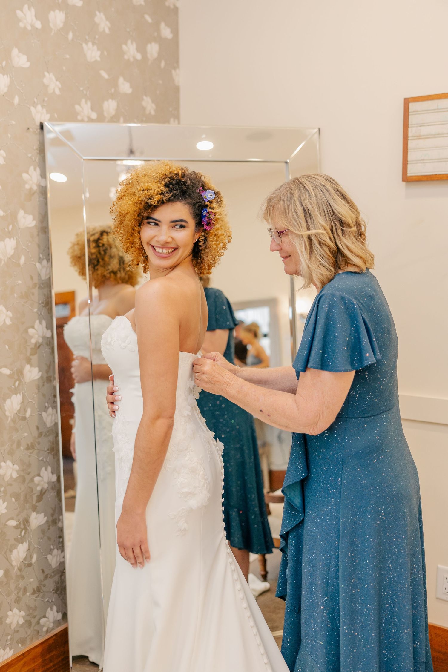 Smiling bride with curly hair in white gown gets dress buttoned by woman in blue dress.