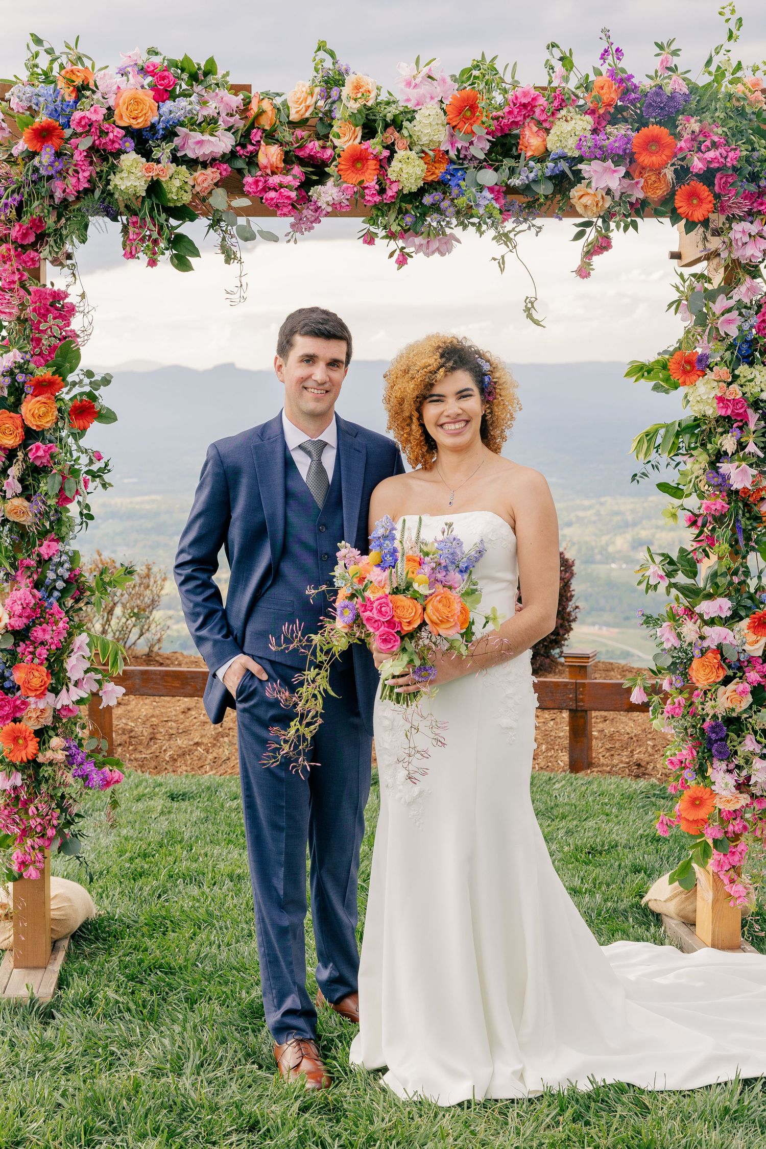 Bride and groom posing under a vibrant floral arch with colorful blooms and mountain views on their wedding day.