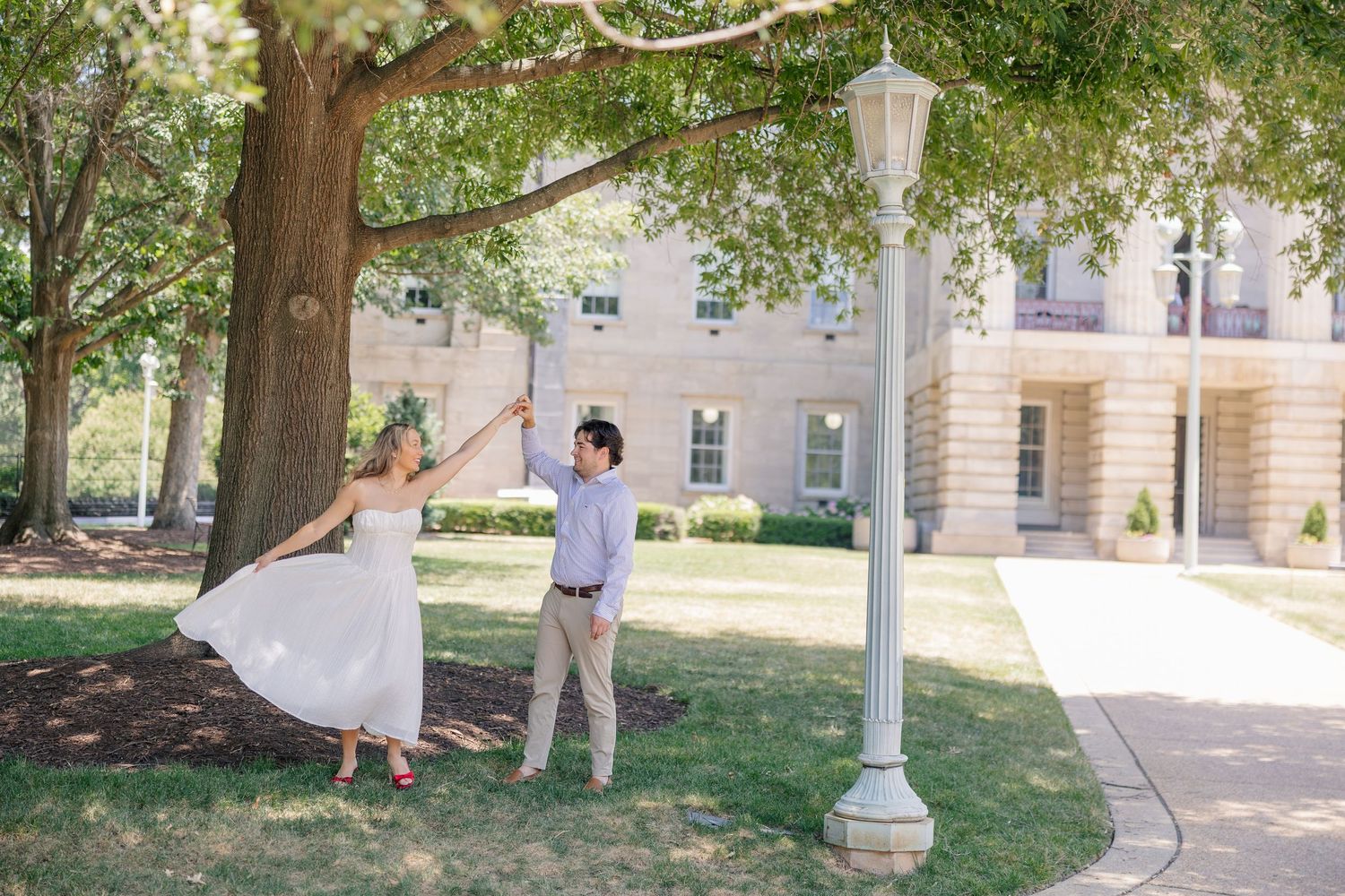 Couple dancing playfully under a large tree near a white lamppost outside a classic brick building.
