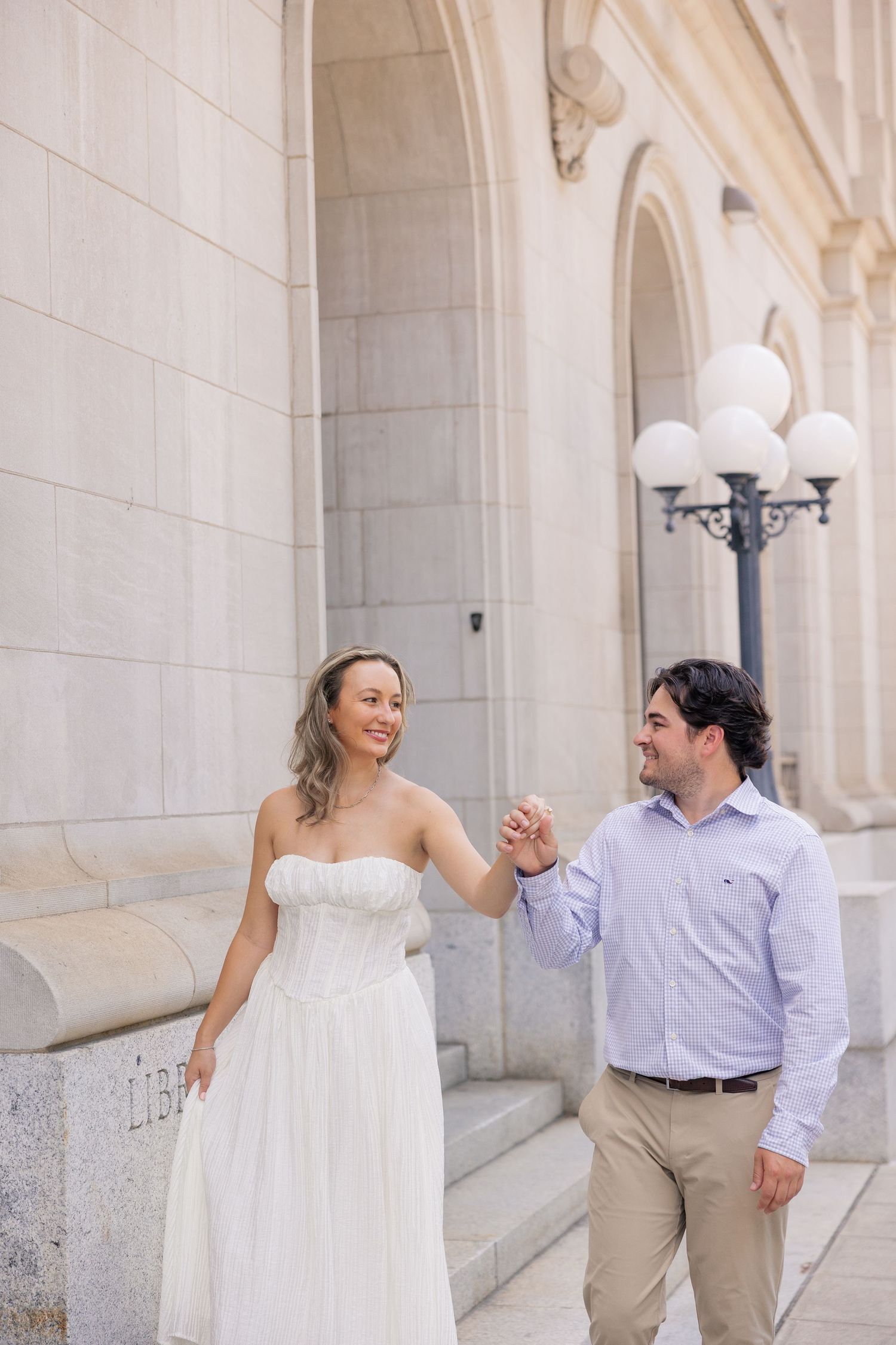 Bride in white strapless gown holds hands with groom in blue shirt walking outside grand stone building.