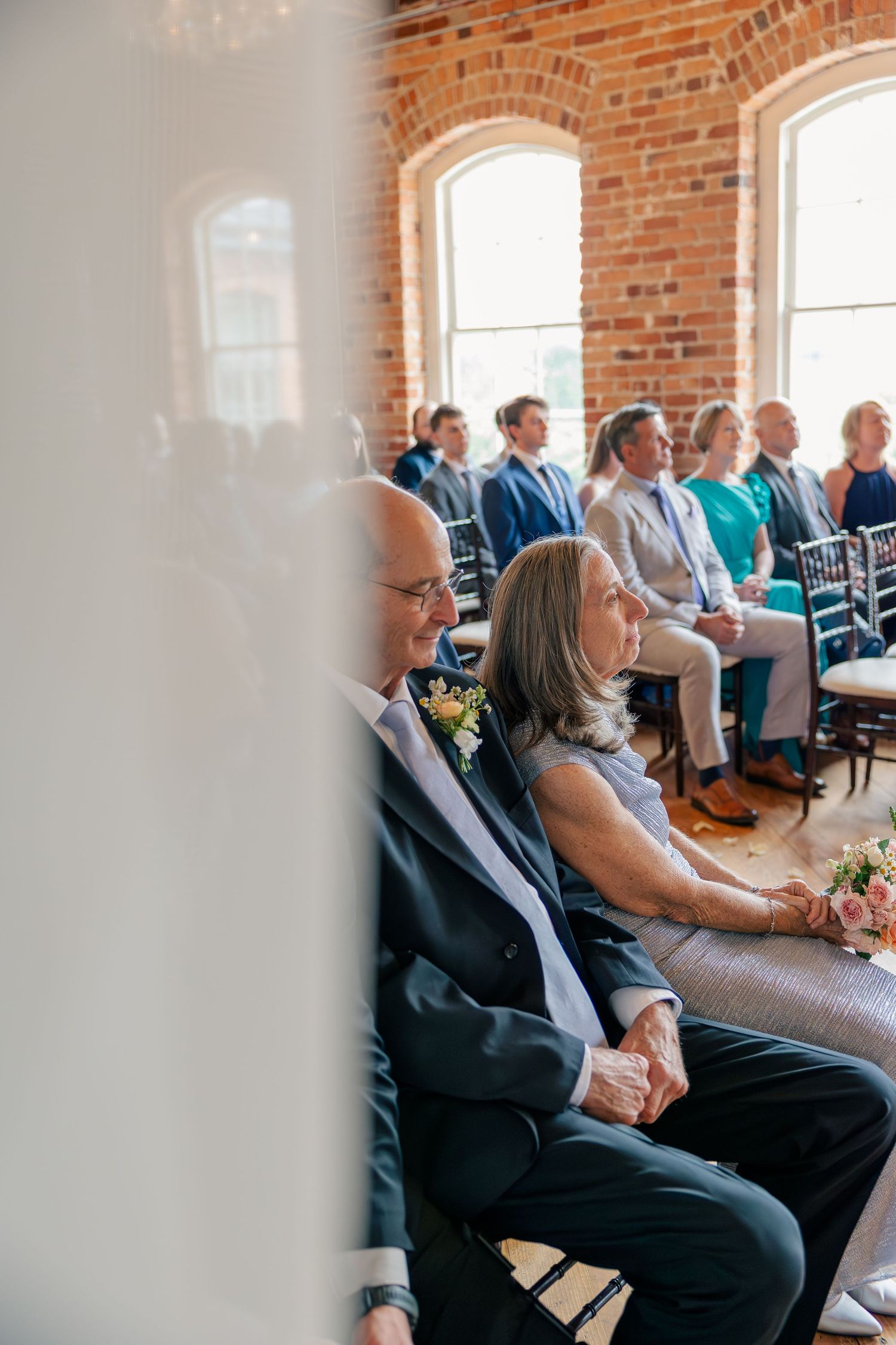 Wedding guests seated in sunlit brick venue hold hands while watching the ceremony unfold.