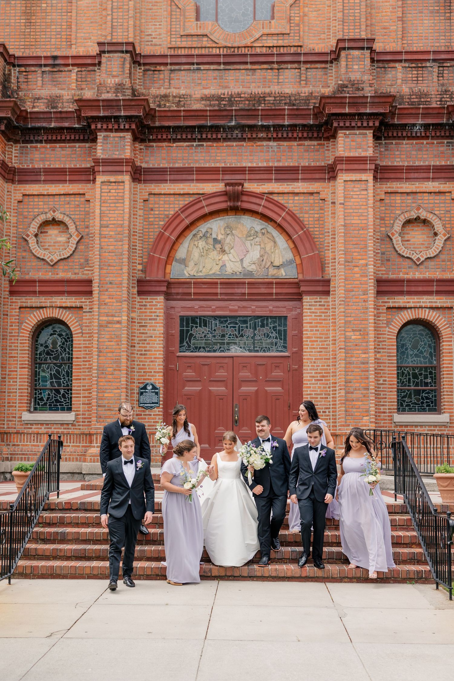 Wedding party descending steps of ornate red brick church with arched doorway and mosaic detail.