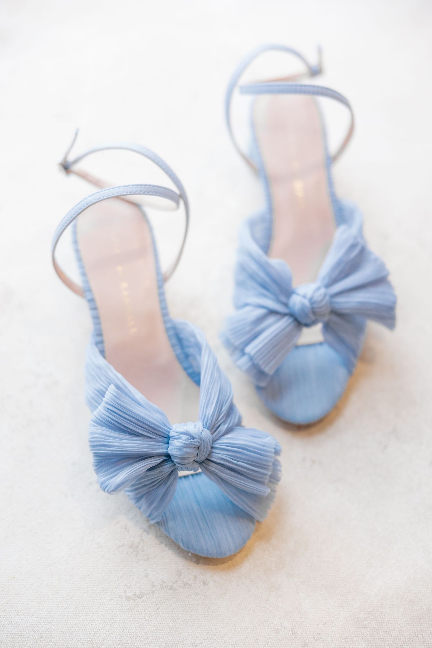 Elegant light blue satin heels with pleated bow detail and ankle strap on a white surface.