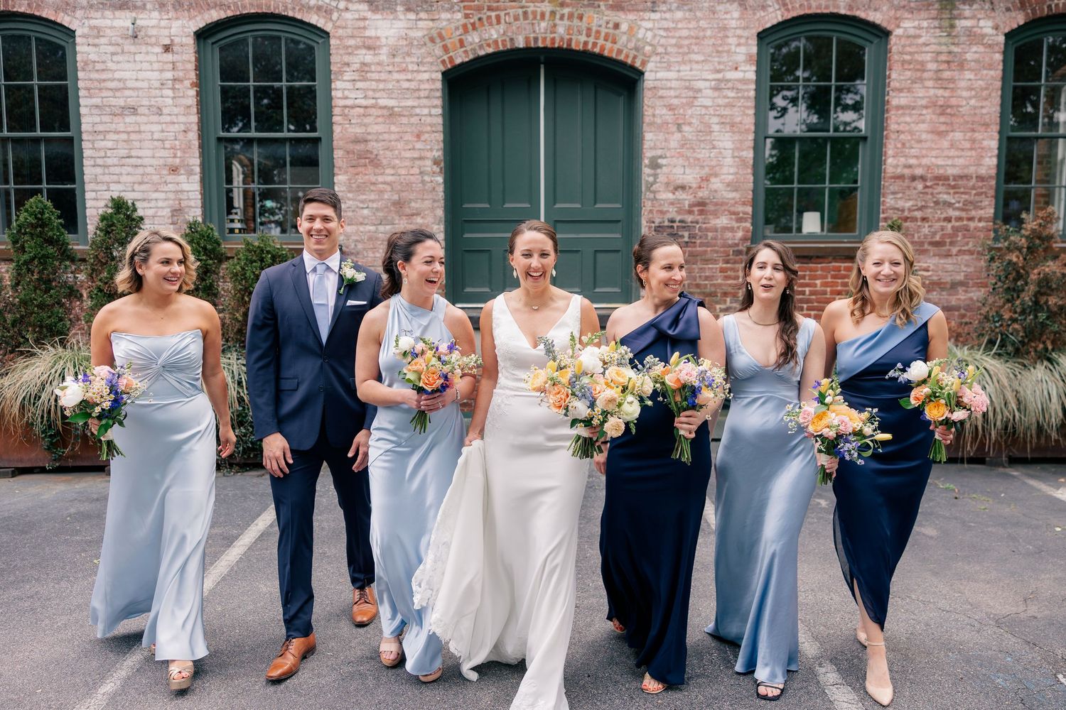 Bride in white gown walks with bridesmaids in blue dresses and groomsman outside brick building.