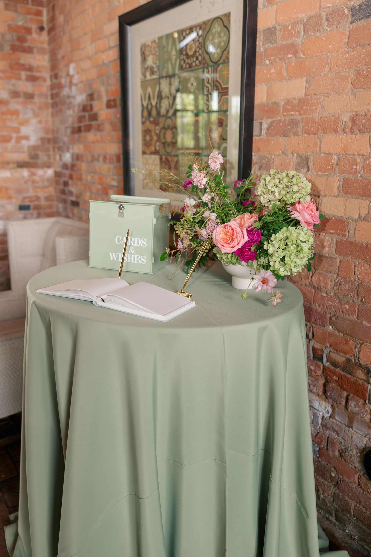 Wedding guest book table with sage green linen, card box, and pink floral arrangement against exposed brick wall.