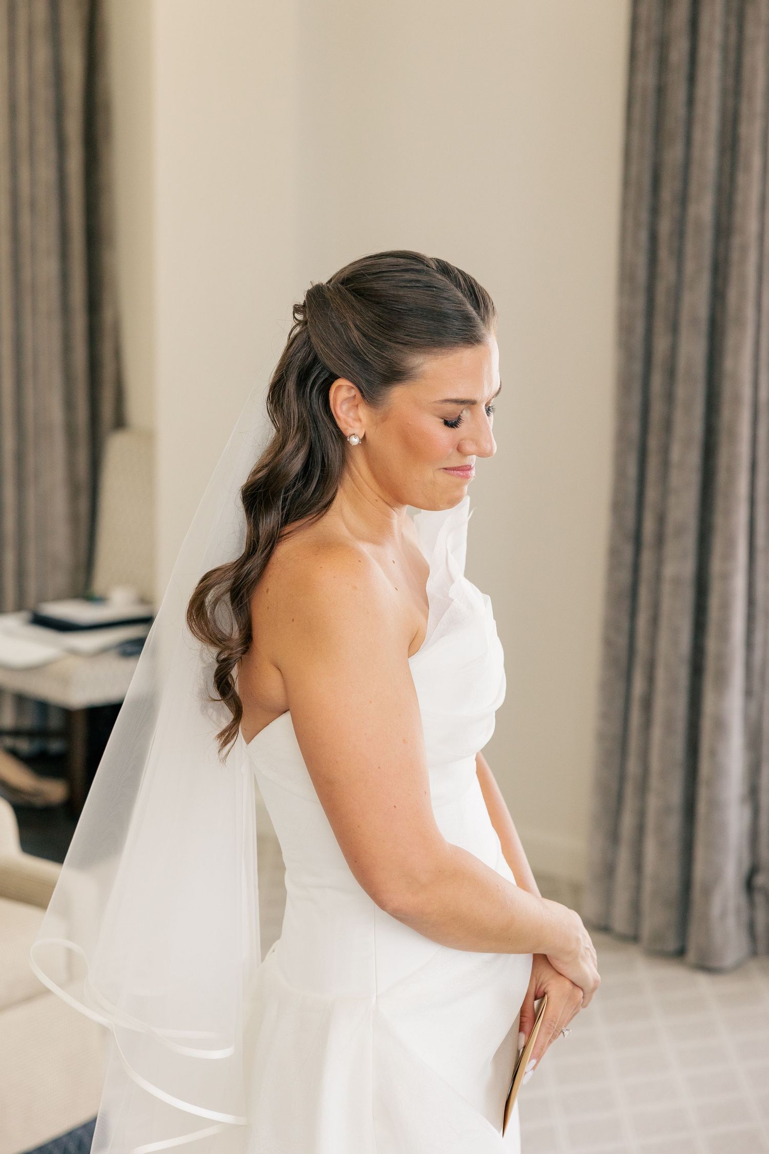 Elegant bride in a strapless white gown and veil poses gracefully in a softly lit room.