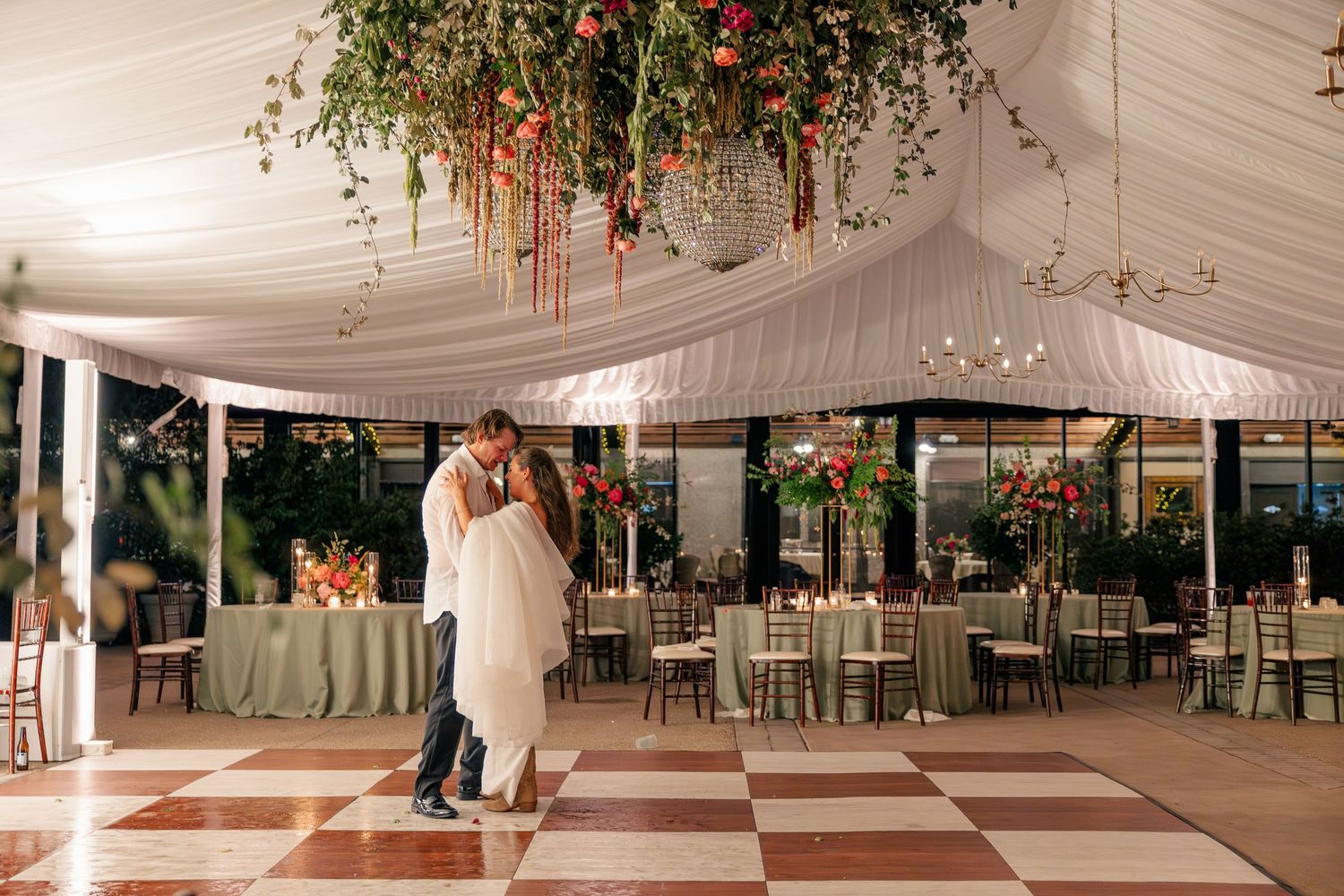 Couple shares first dance under floral ceiling installation in elegant tented wedding reception with checkered dance floor.