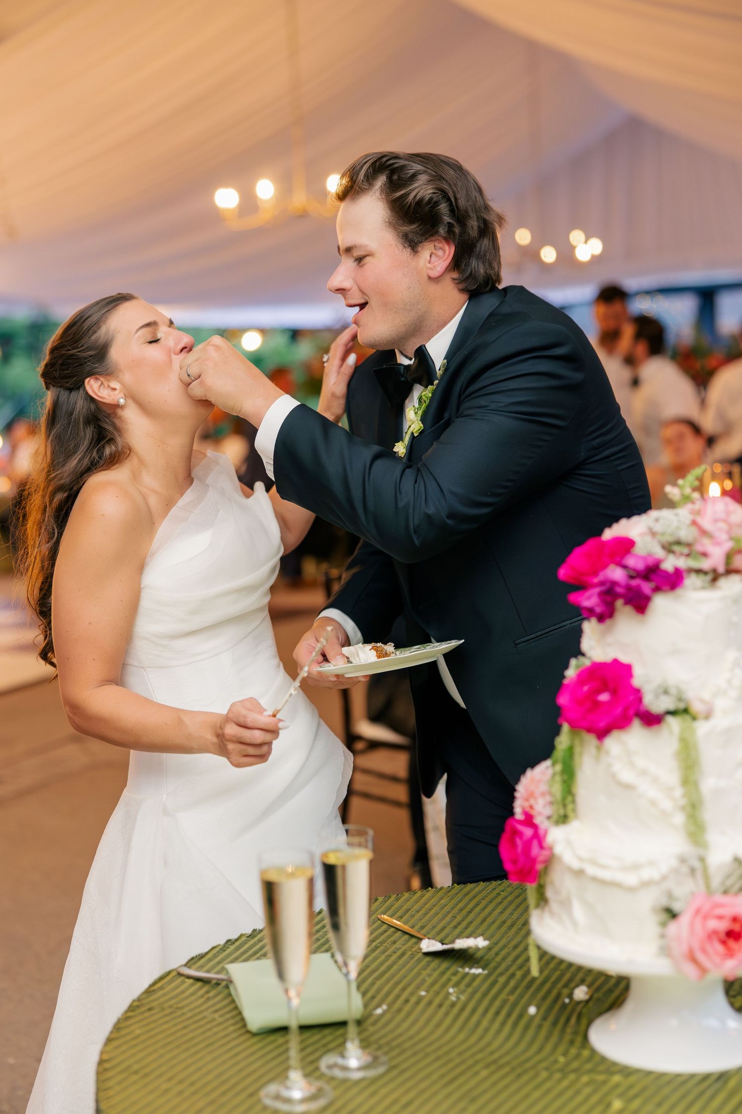 Bride and groom laugh while feeding each other wedding cake at elegant tented reception.