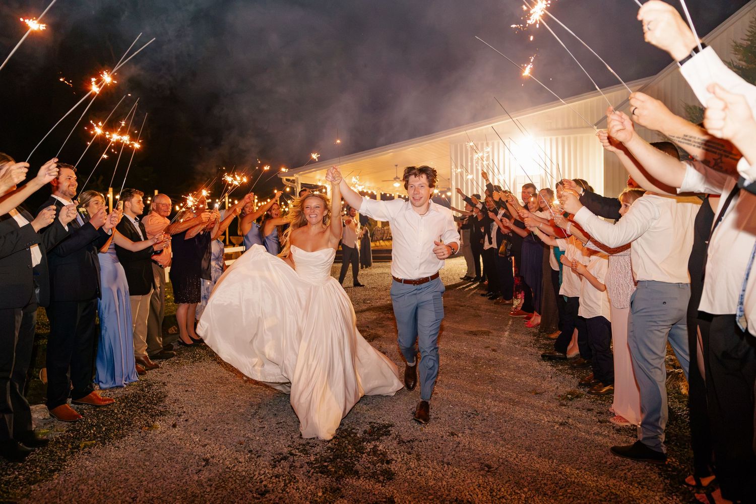 Bride and groom run through sparkler exit tunnel at night outside illuminated wedding tent.