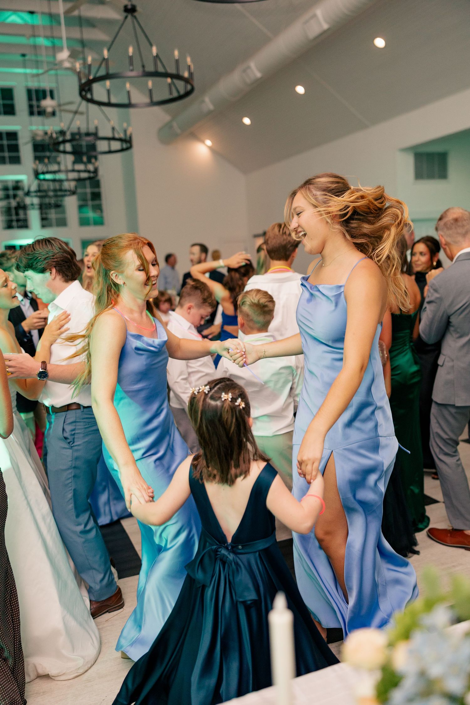Bridesmaids in blue dresses dance joyfully with flower girl at lively indoor wedding reception.