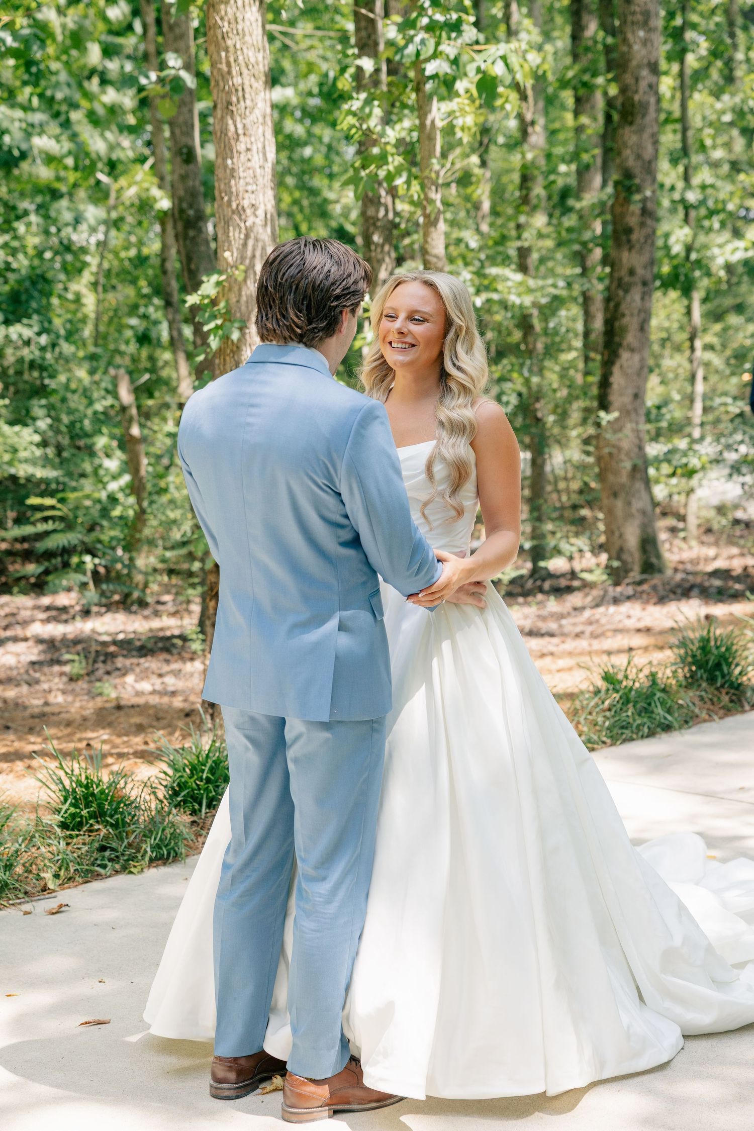 Bride in white ball gown smiles at groom in light blue suit during outdoor first look among trees.