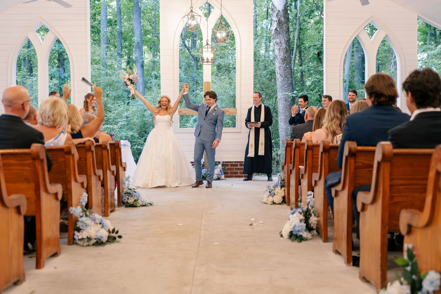 Joyful newlyweds raise arms in celebration at altar of stunning glass chapel surrounded by trees.