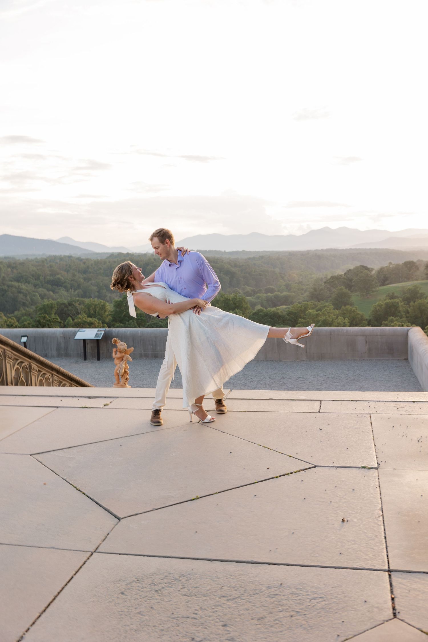A couple in white and lavender outfits dancing on a rooftop terrace with a scenic mountain view at sunset.