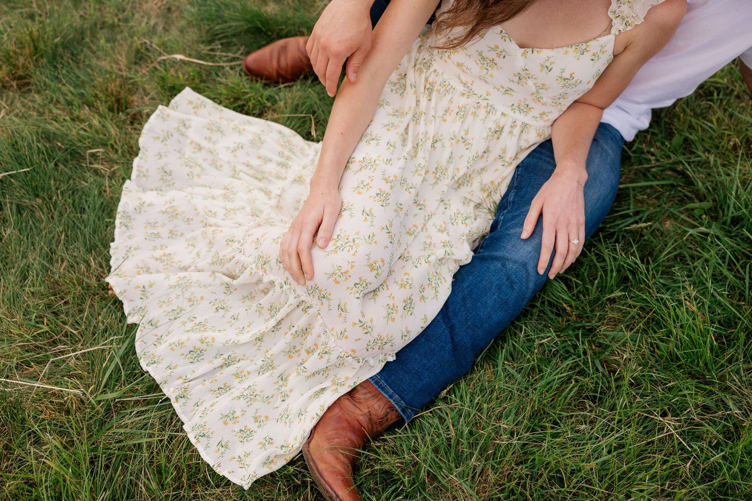 A floral dress and denim-clad couple sitting together on grassy ground with brown boots visible.