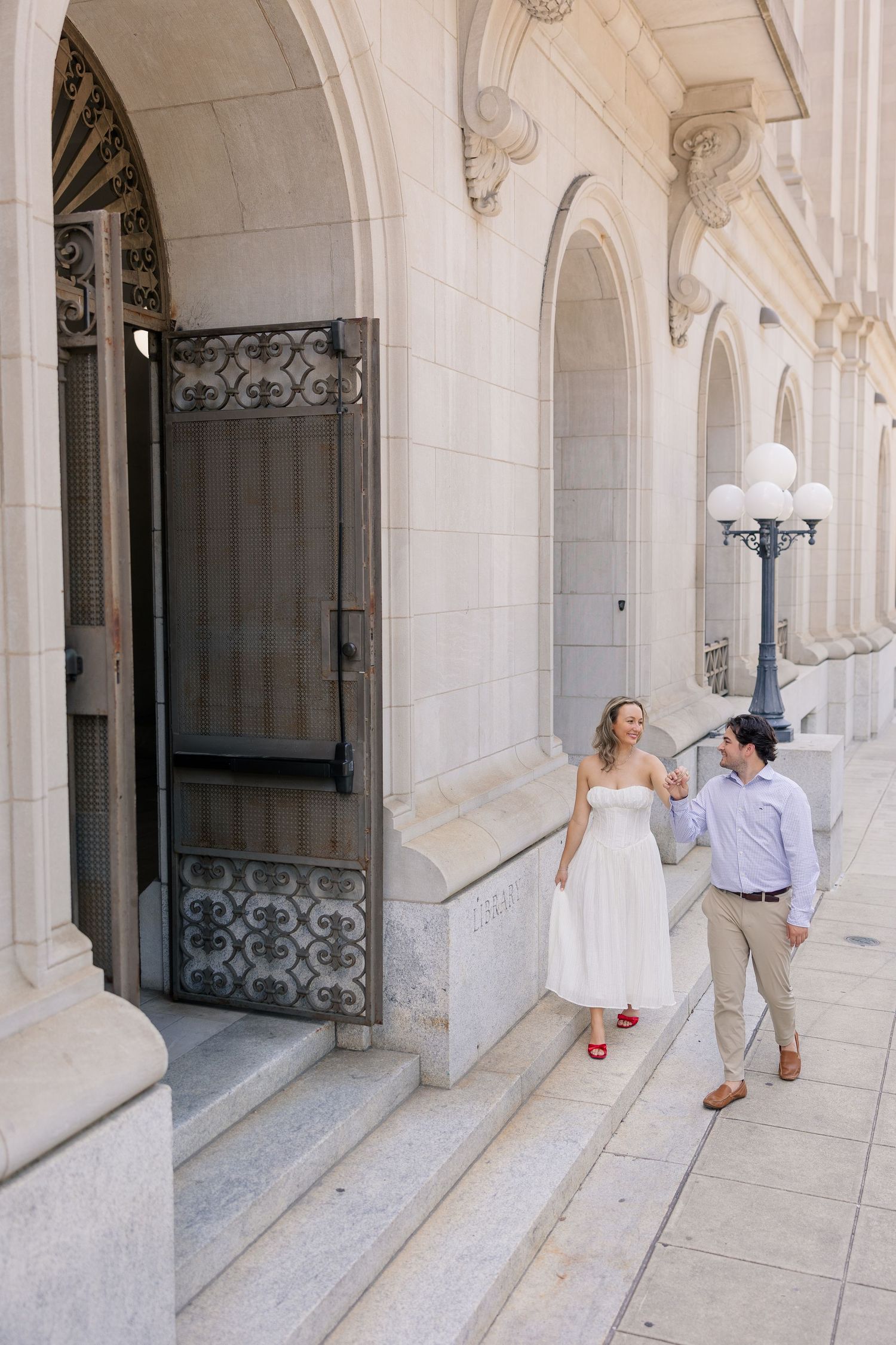 Engaged couple walking and laughing together on steps of elegant white stone building with ornate iron doors.