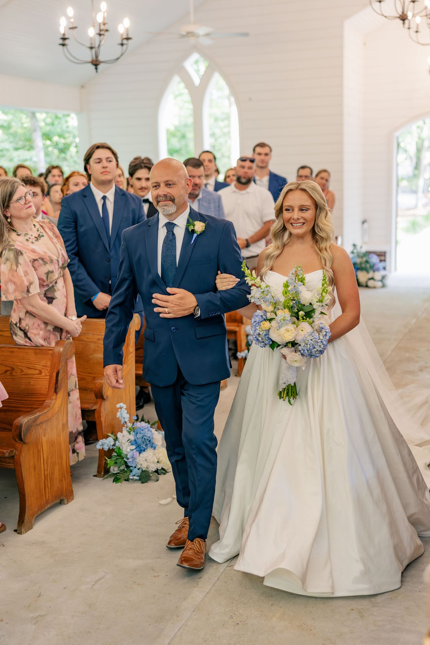 Bride in white ball gown walking down church aisle with her father during wedding ceremony.