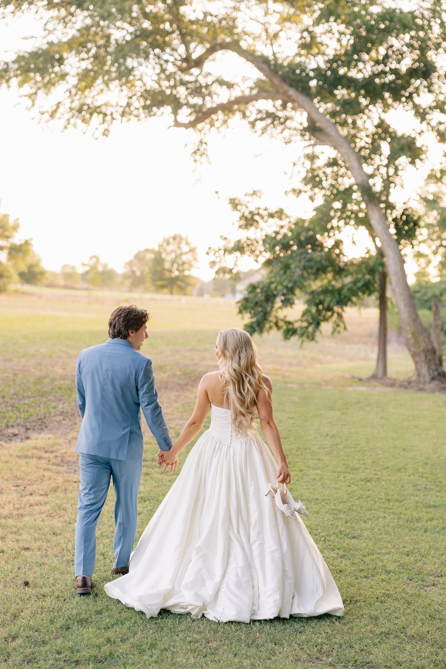 Bride and groom walk hand in hand through a sunlit green field at golden hour.