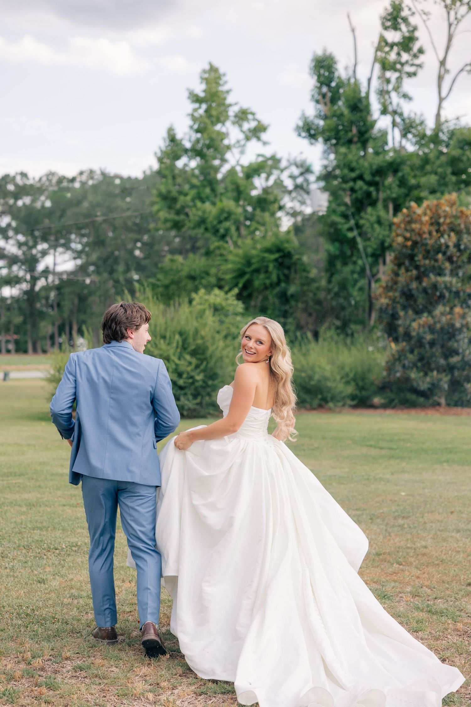 Bride in strapless white ball gown walks with groom in blue suit across lush green lawn.