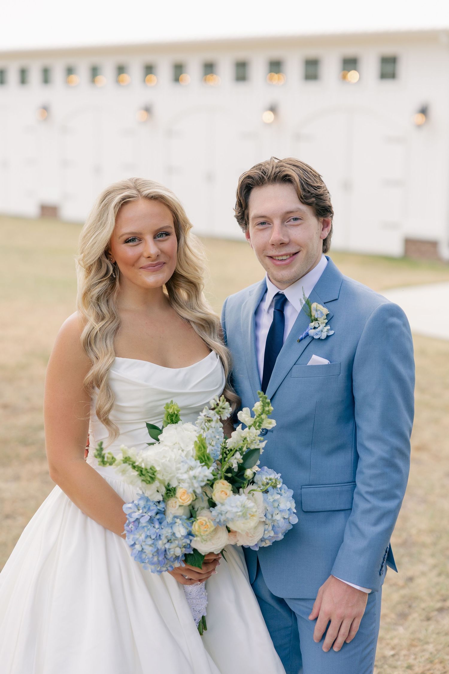 Bride and groom posing together outdoors, bride holding blue and white floral bouquet in front of white barn.