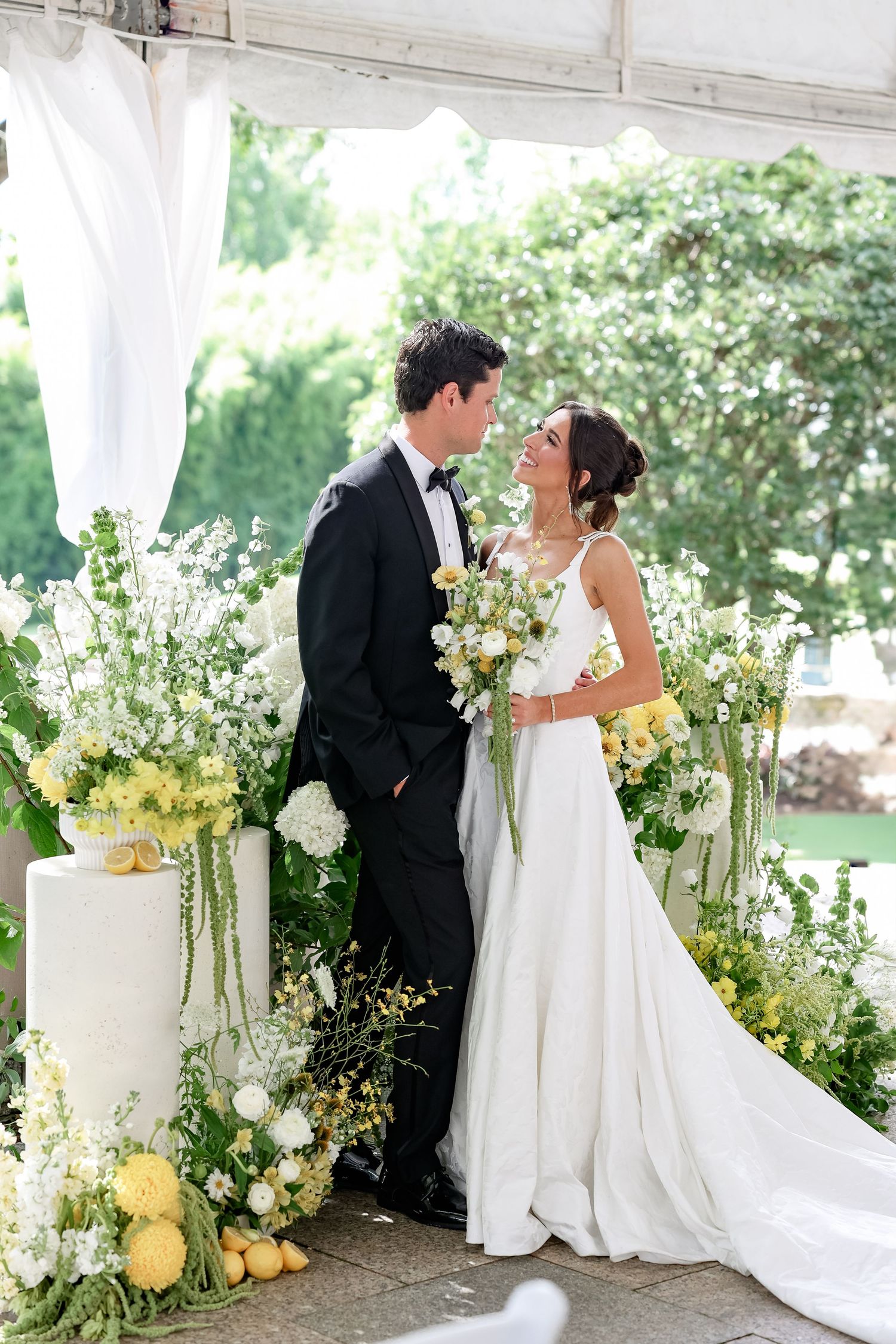 Bride and groom share a romantic moment at floral altar with yellow and white flowers during outdoor wedding ceremony.