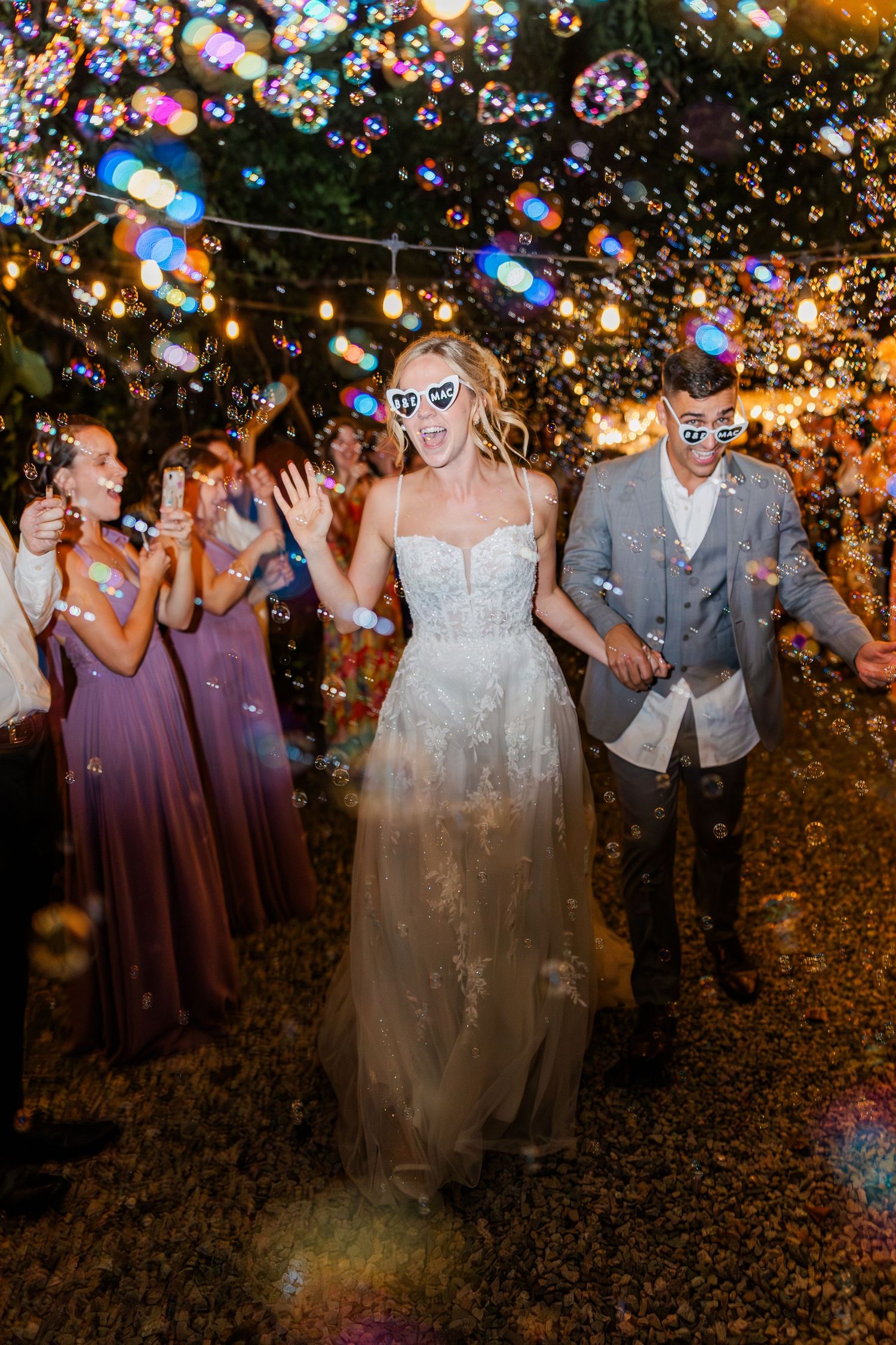 Bride and groom wearing fun masks walk through bubbles and confetti at their outdoor evening wedding reception.