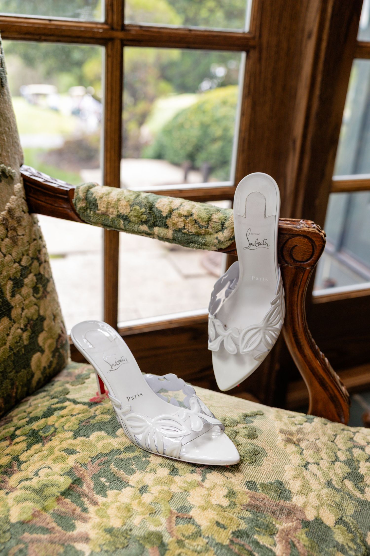 White Christian Louboutin bridal heels with floral details displayed on a floral upholstered chair near a window.