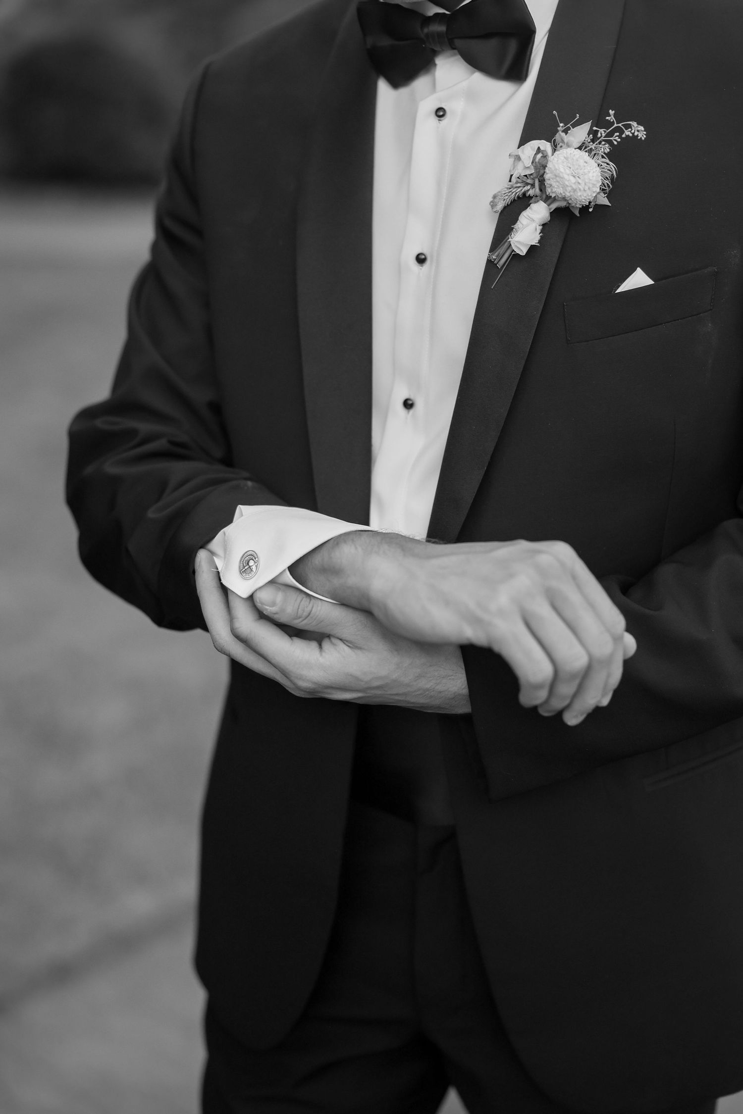 Black and white photo of a groom in a tuxedo adjusting his cufflinks, wearing a boutonniere and bow tie.