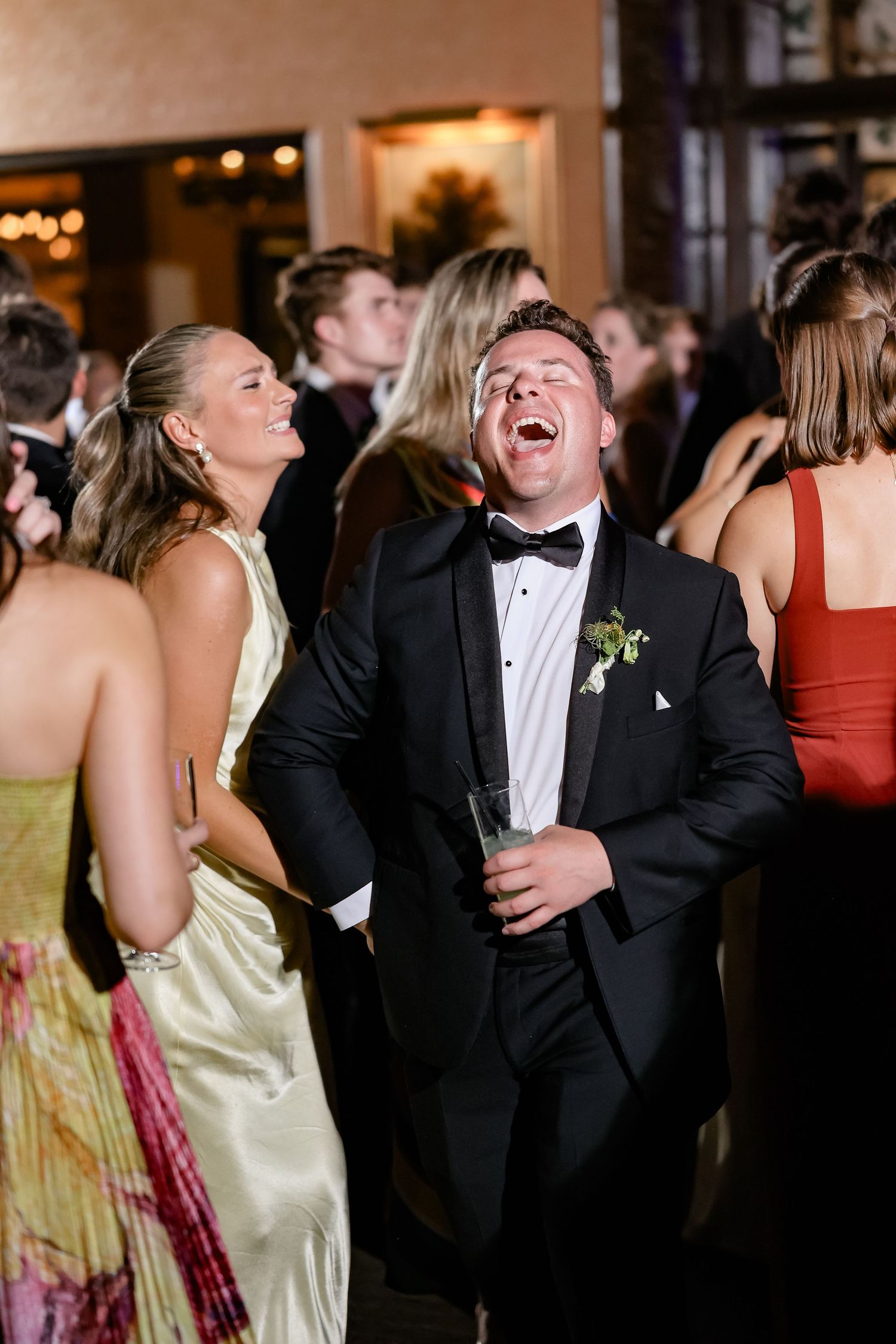Wedding guests in formal attire laugh and celebrate joyfully on the dance floor at a reception.