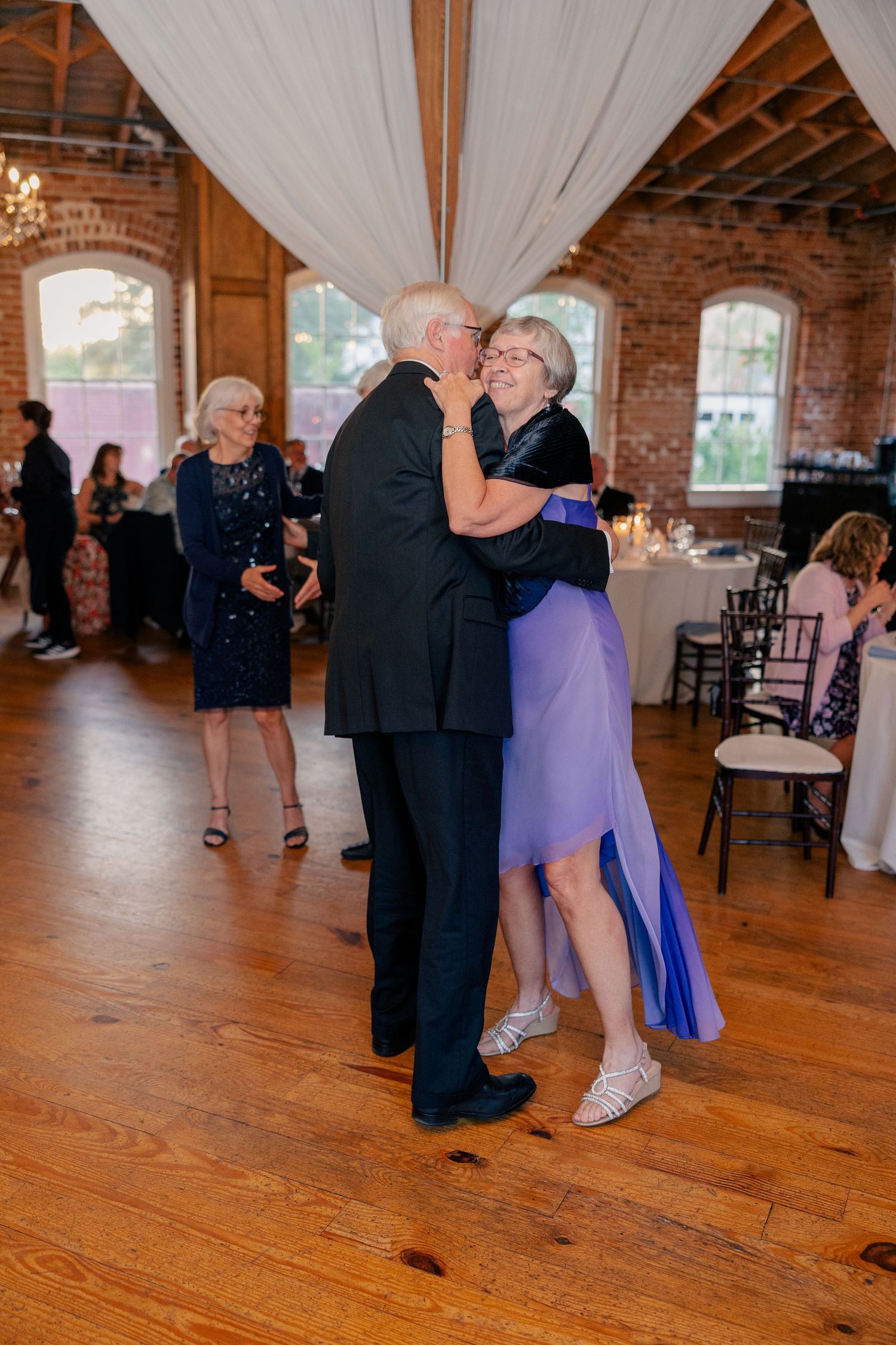 Elderly couple shares a romantic dance together at wedding reception in rustic brick ballroom.