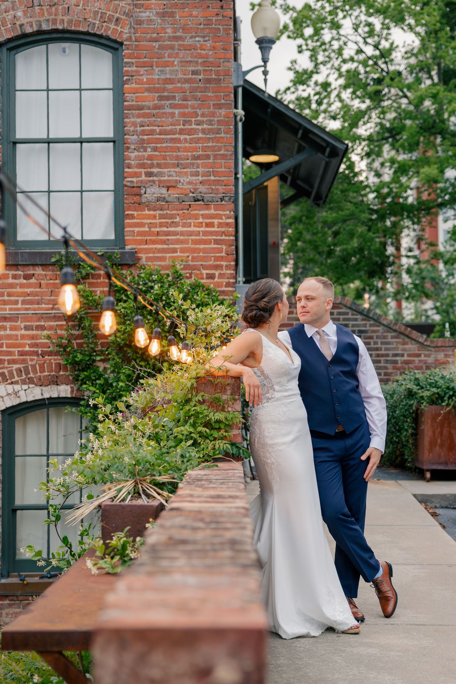 Bride and groom pose romantically outside a charming brick building with string lights and lush greenery.