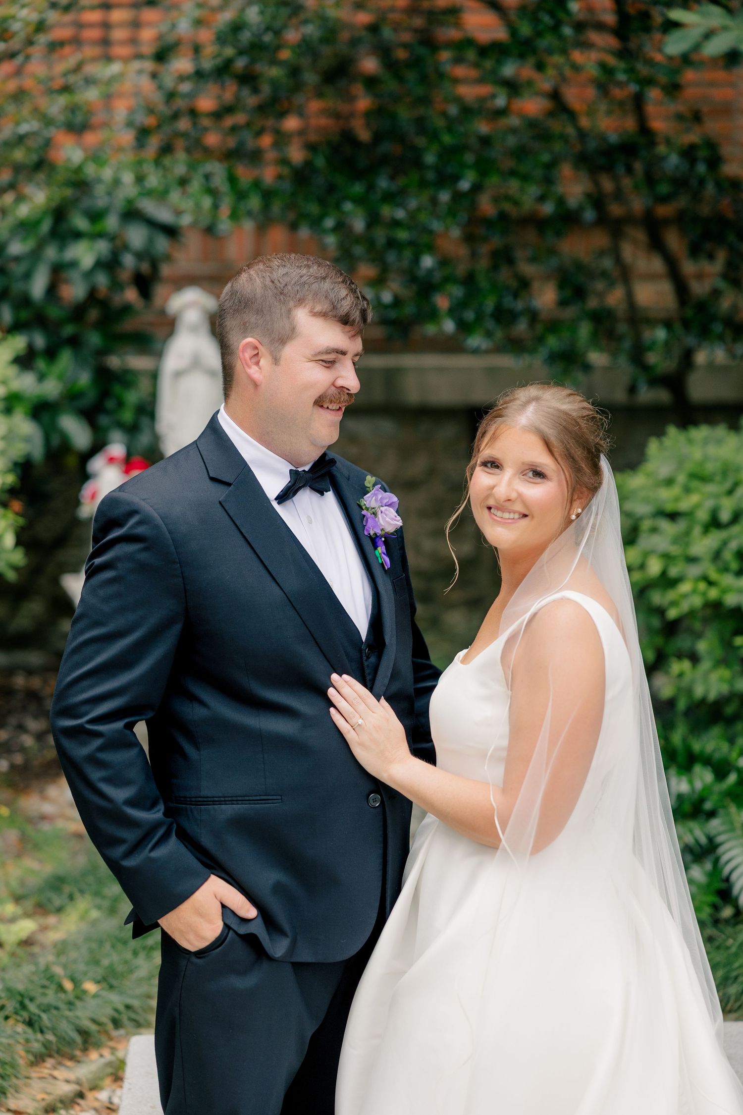 Bride in white gown smiling at camera while groom in black tuxedo looks at her outdoors.