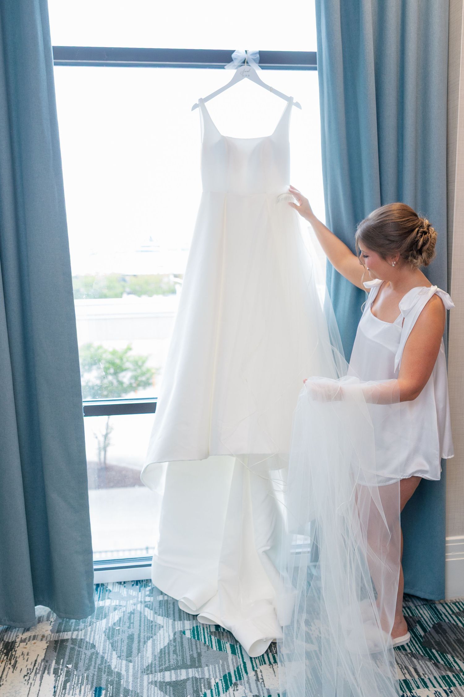 Bride in white robe admiring her elegant wedding gown hanging by a bright hotel window before the ceremony.