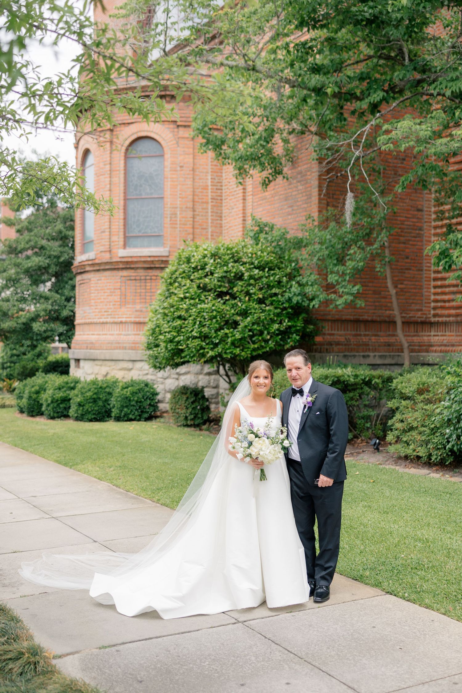 Bride and groom portrait outside historic brick church with bouquet and flowing veil.