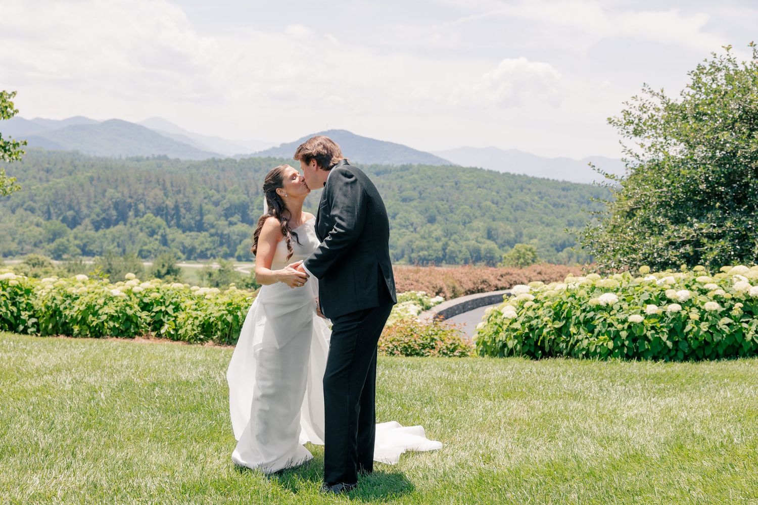 Bride and groom share first kiss on lush green lawn with mountain views and blooming gardens at outdoor wedding.