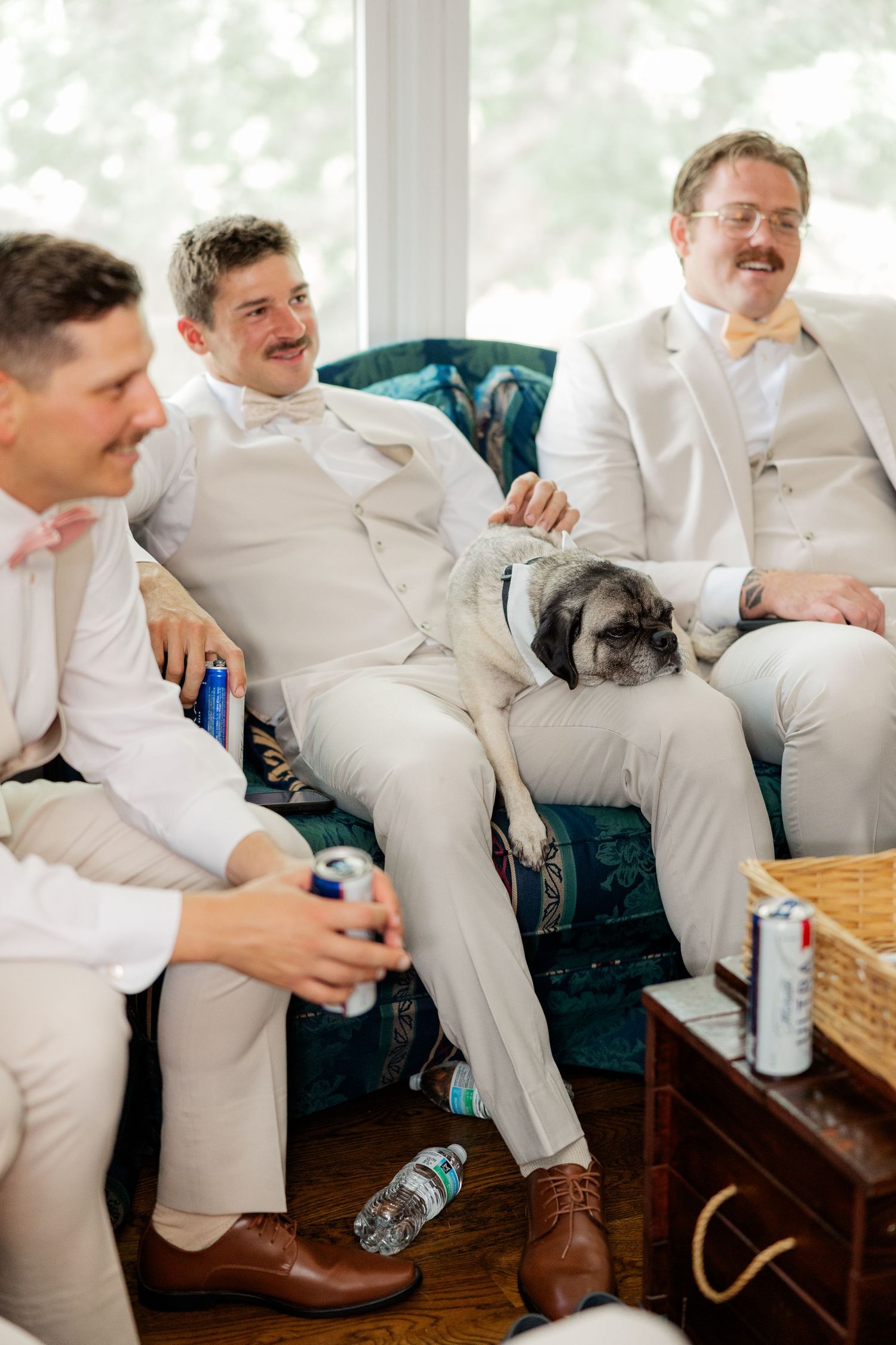 Groomsmen in white suits share laughs while putting on brown leather dress shoes before wedding ceremony.
