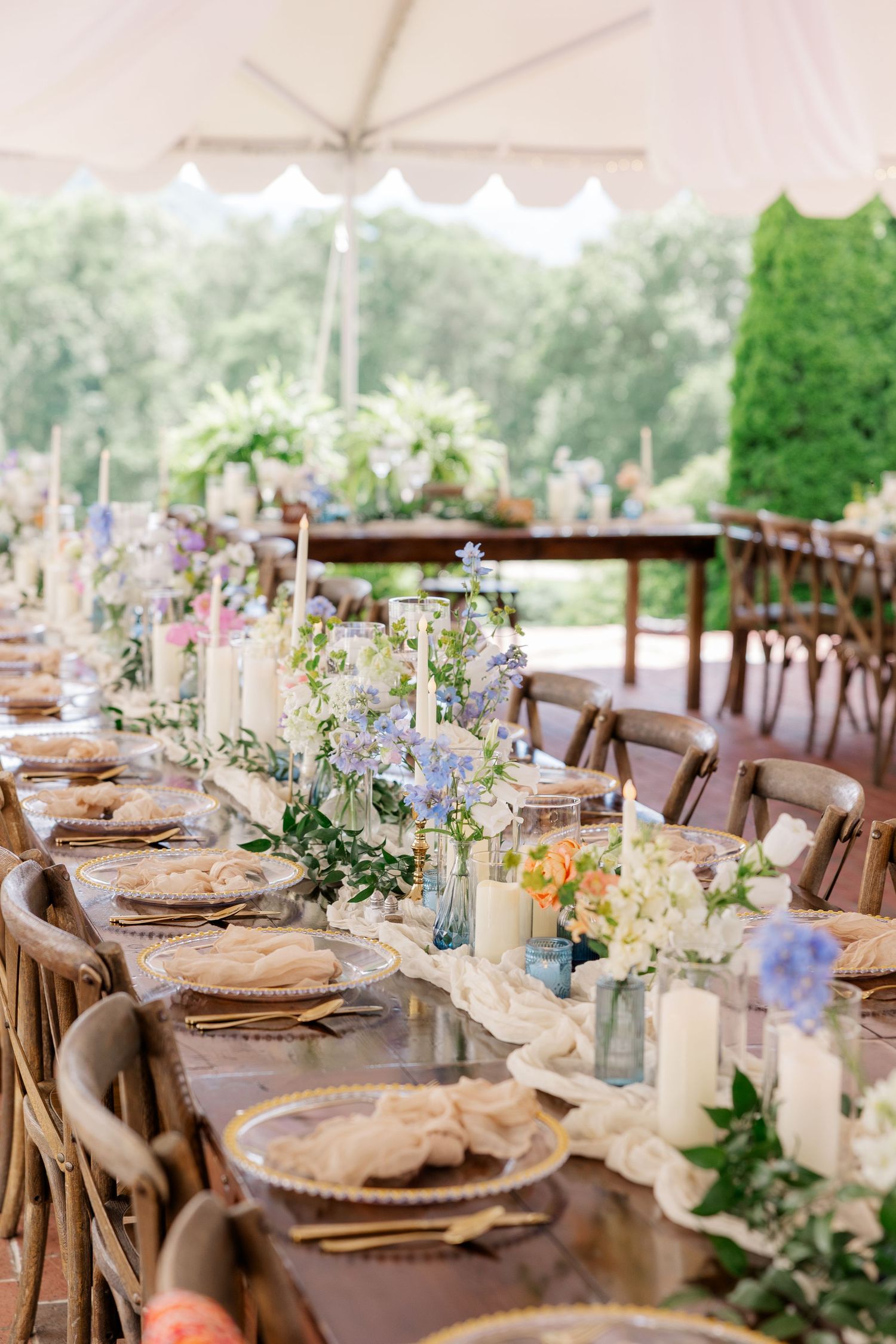 Elegant wedding reception table settings with delicate floral centerpieces and rustic wooden chairs under a white tent.