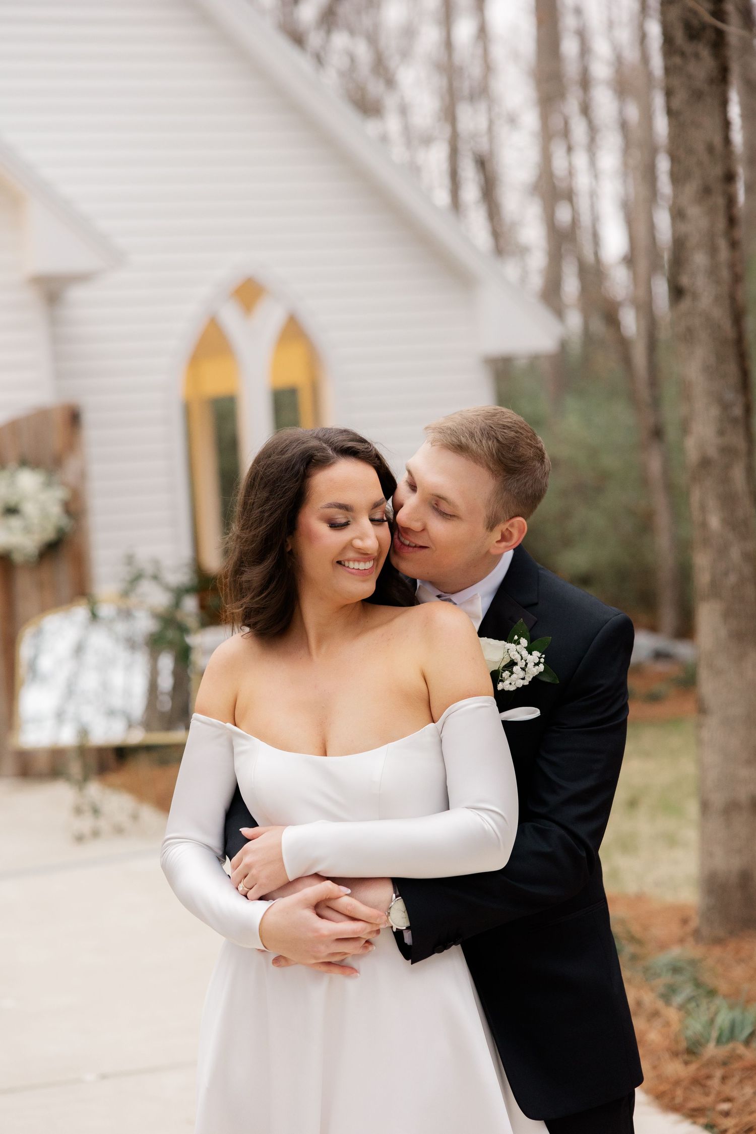 A romantic wedding couple embraces in front of a white chapel with autumn trees in the background.