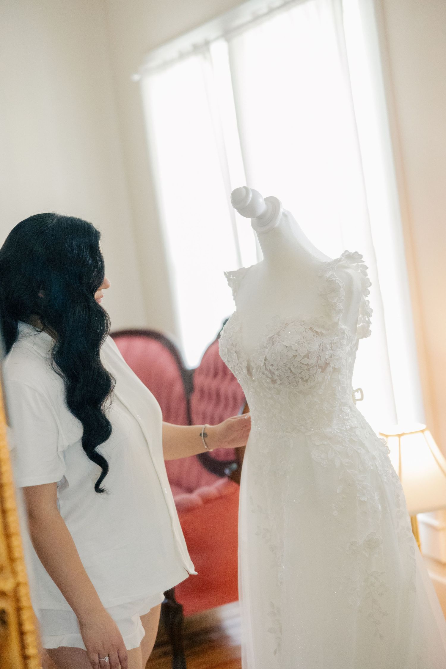 A white lace wedding dress hanging in soft window light shows intricate details of the bodice and train.