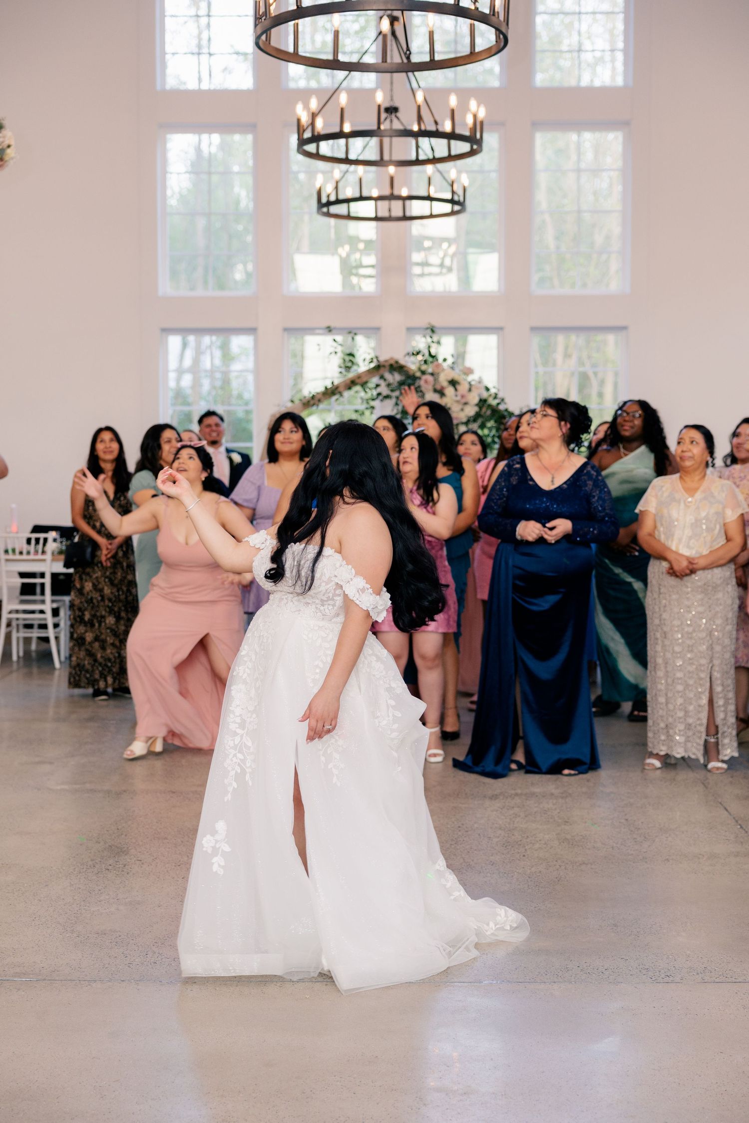 Wedding reception scene showing guests dancing beneath elegant circular chandeliers in a bright white venue.