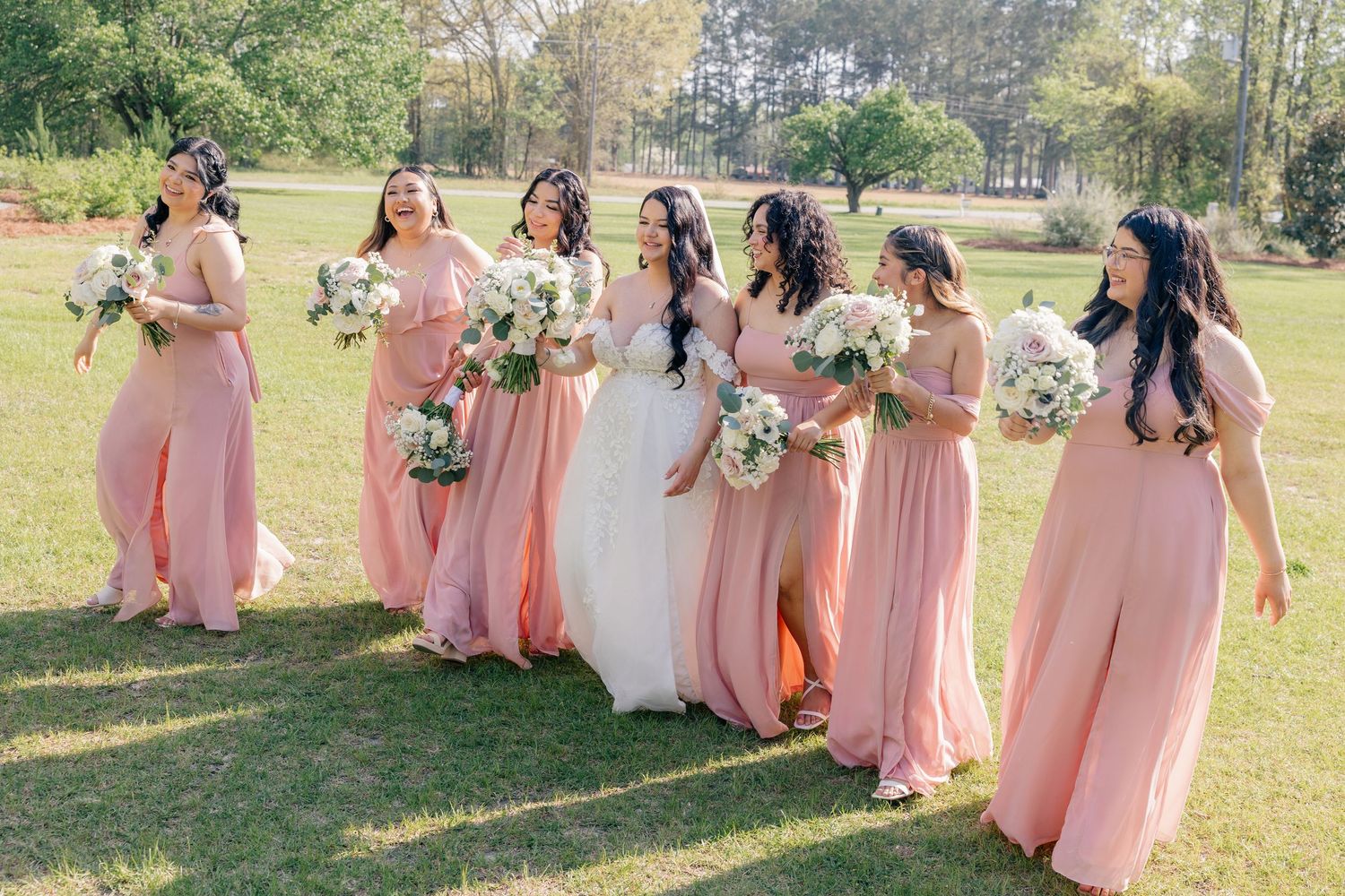 Bridesmaids in coral pink dresses pose with the bride on a sunny lawn.