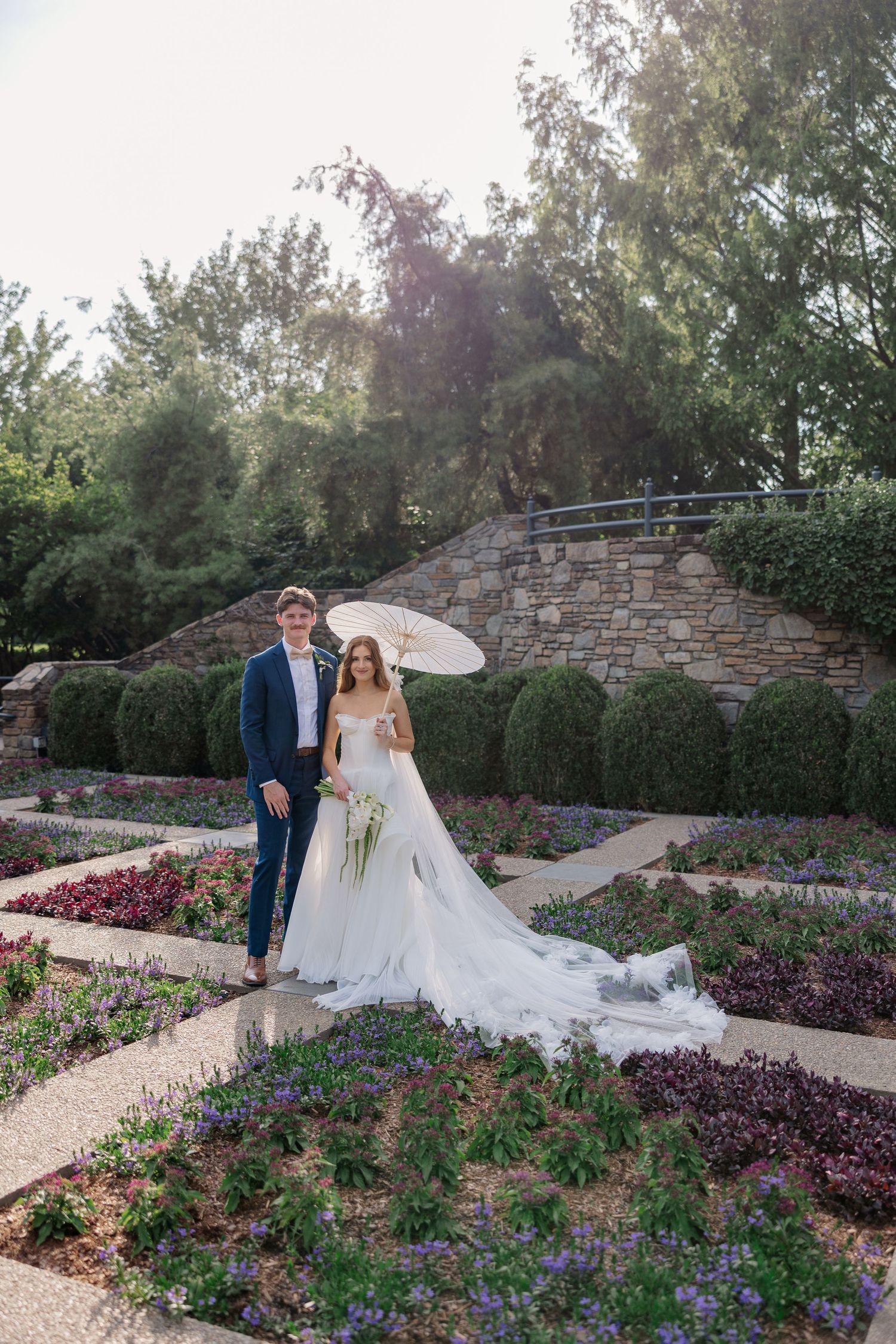 Wedding couple standing in a garden with stone wall and purple flowers.
