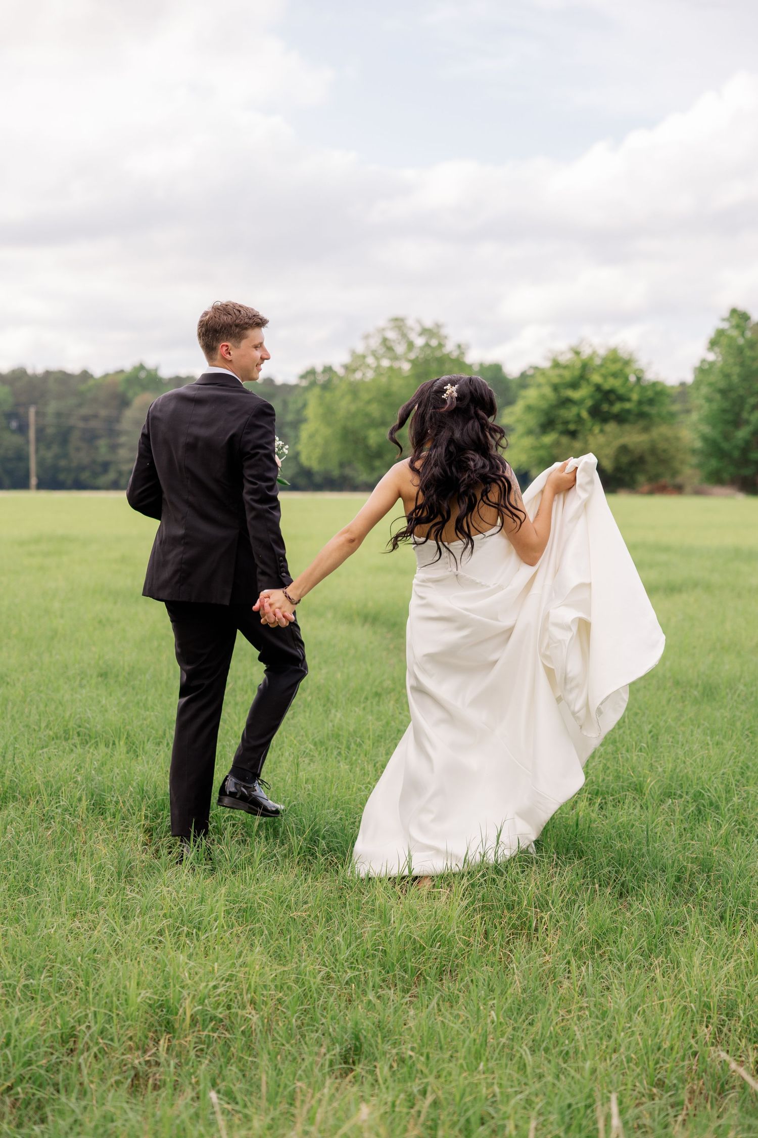 Wedding couple walks through grassy field while holding hands and lifting dress fabric.