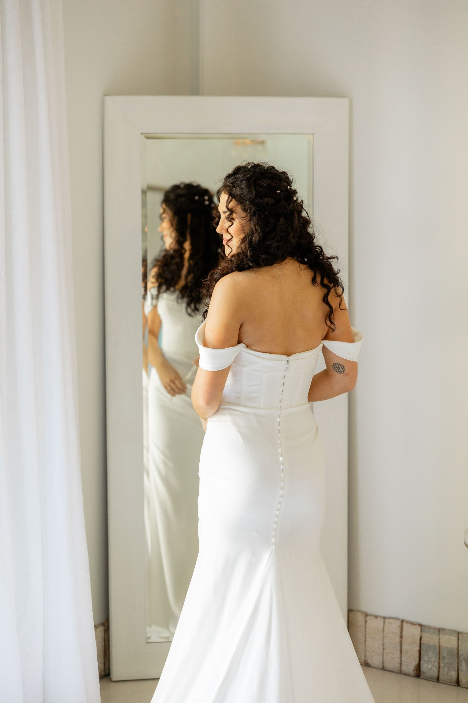 A bride in a fitted off-shoulder white wedding gown looks at her reflection in a full-length mirror.