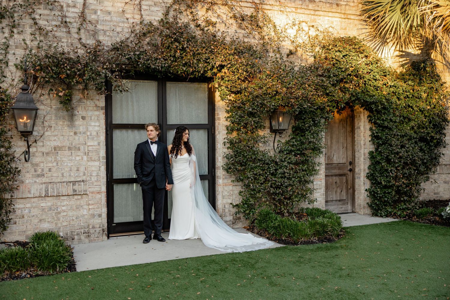 Couple poses in formalwear against stone building covered in lush green vines.